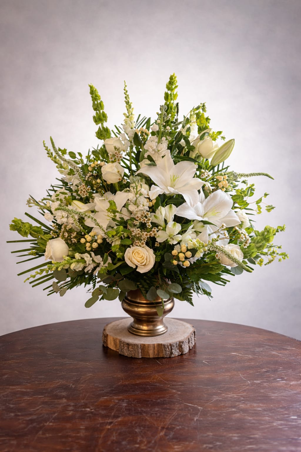 Traditional white urn - A serene, classic display of white lilies and roses makes this flower arrangement a graceful choice for sympathy or a formal wedding centerpiece. The Traditional white urn arrangement delivers a calm, elegant presence-ideal for honoring someone dear or marking a solemn, refined occasion-so the person you're remembering will feel respected and truly seen.  This piece features large, airy white lilies, creamy white roses and tall snapdragon spikes arranged in a rounded, expansive form. Small white filler blooms and pale berry clusters add delicate texture, while layered greenery gives the design a full, structured silhouette. The flowers are presented in a metallic gold vase set on a natural wood slice base, combining formal white-and-green styling with a warm, grounded accent.  Perfect for a funeral service, memorial table, or as a wedding altar arrangement, this composition reads as both traditional and quietly modern. Its generous scale and classic palette make it easy to place in chapels, reception rooms, or family homes; to keep it looking fresh, change the water regularly and trim stems as needed.