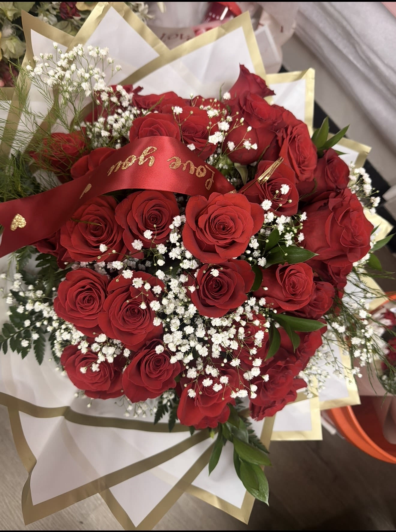 Bouquet of red roses and white filler flowers wrapped in white and gold paper with a red love message ribbon.