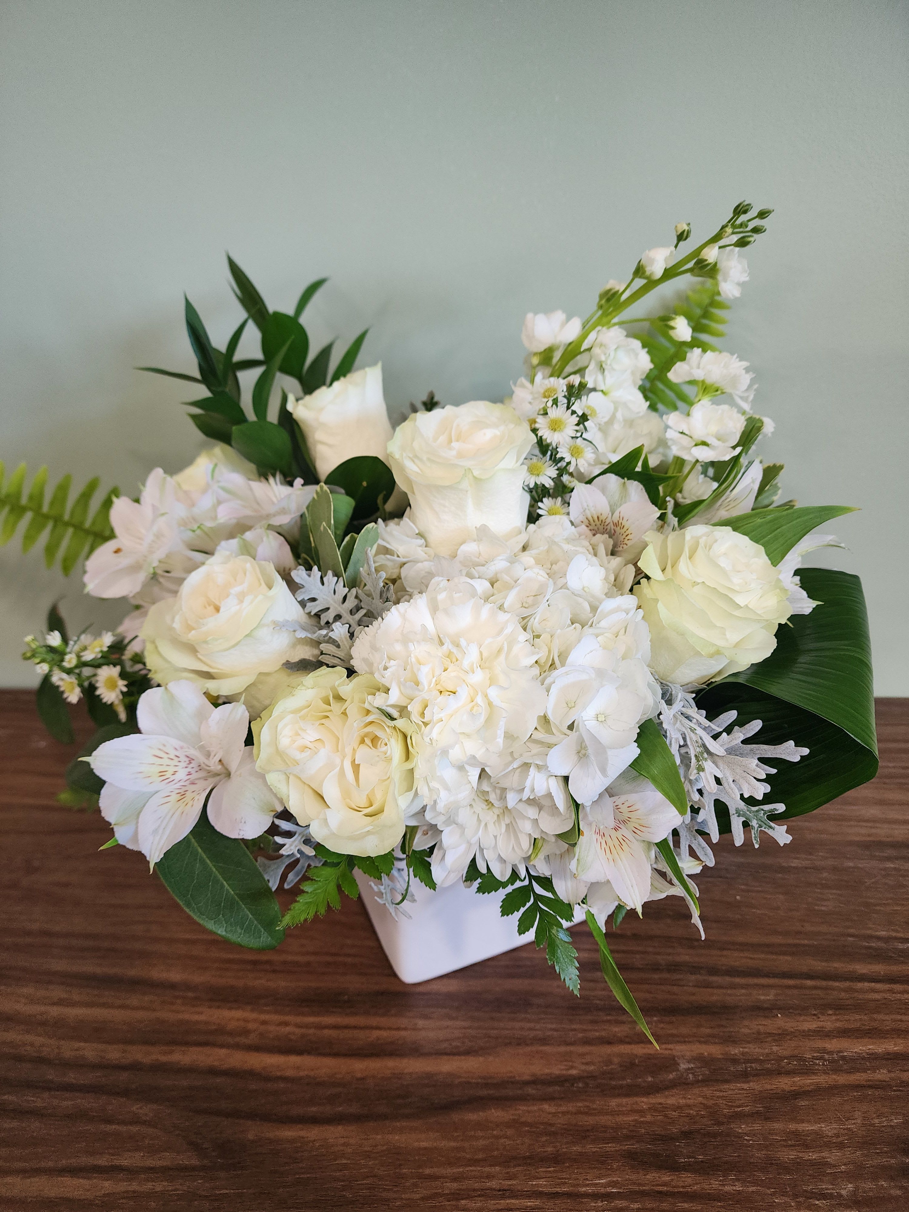 Graceful Blooms by MG Florist  - Graceful Blooms by The Magic Garden Florist, Rochester, NY  Bring timeless beauty and serene elegance into any space with our stunning White Floral Cube arrangement from Magic Garden Florist. Designed in a modern white ceramic cube, this sophisticated piece features a harmonious blend of fresh white hydrangeas, classic white roses, and delicate white alstroemeria, and carnations.  Perfect for weddings, sympathy occasions, anniversaries, or minimalist home décor, this all-white floral arrangement symbolizes purity, grace, and tranquility. Each bloom is carefully selected and artistically arranged by expert florists to create a balanced, lush presentation that complements both contemporary and traditional settings.  Key Features:  Premium fresh white hydrangeas, roses, and snapdragons Stylish white ceramic cube vase (perfect for reuse) Handcrafted floral design for a luxurious, high-end look Ideal for gifts, events, centerpieces, and home décor  Whether you're searching for a “white flower arrangement delivery,” “elegant sympathy flowers,” or a “modern floral centerpiece,” this exquisite design is the perfect choice. Order today from Magic Garden Florist for a refined floral experience delivered with care.
