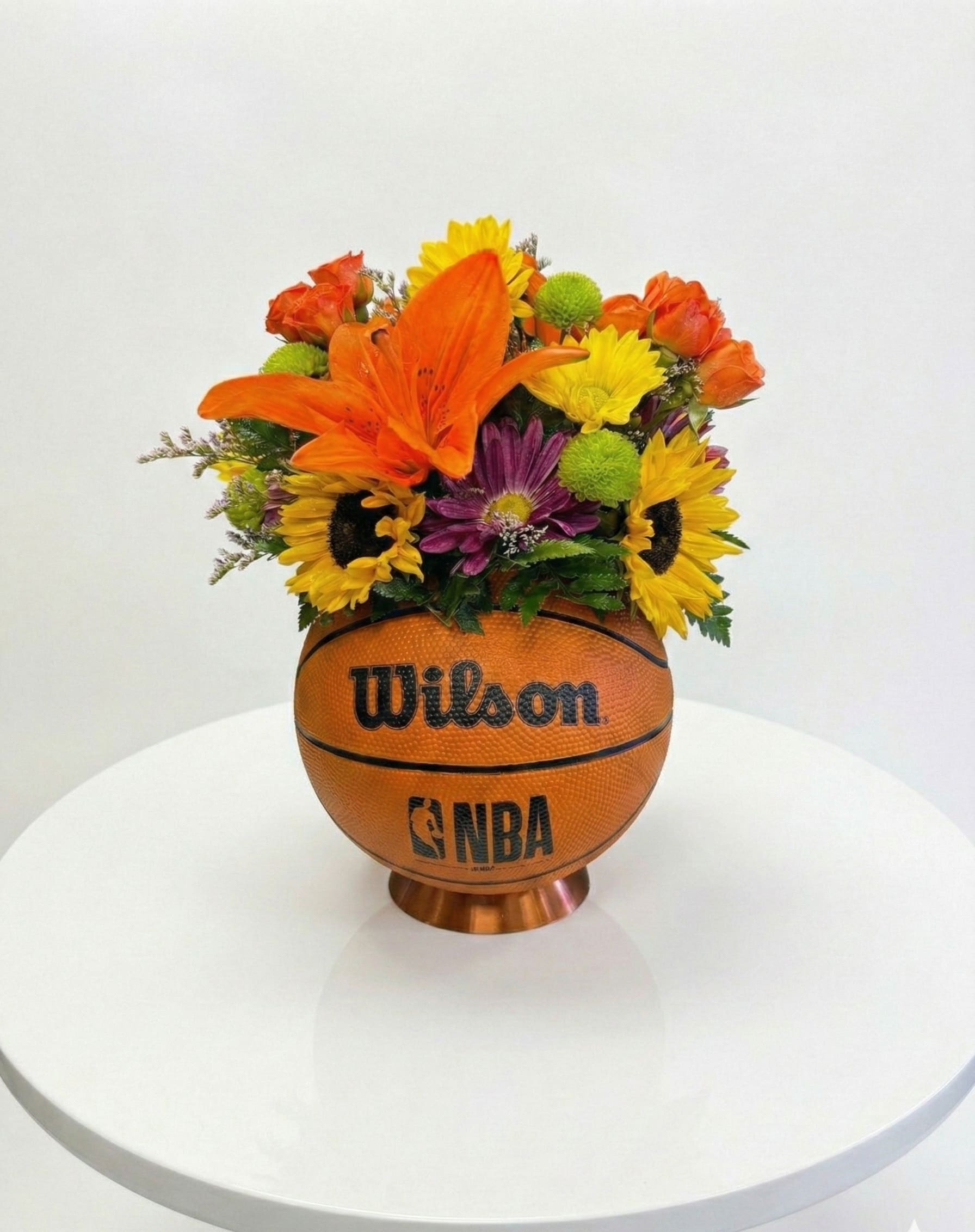 NBA All-Star Tribute - Where Courtside Meets Countryside.  Elevate your gifting game with The All-Star Tribute, a one-of-a-kind floral experience designed for the ultimate basketball enthusiast. This isn’t just a bouquet; it’s a championship-caliber centerpiece that bridges the gap between rugged athleticism and refined botanical art.  Nestled inside a genuine, textured Wilson mini NBA basketball, this arrangement features a vibrant "starting lineup" of premium blooms:  Hero Lilies: Bold, fiery orange lilies that command attention from every angle.  Sun-Kissed Sunflowers: Deep yellow petals that bring a radiant, high-energy glow.  Textural Accents: A mix of purple chrysanthemums and "button" poms for a sophisticated, layered look.  Perfect For:  Celebrating a big win or a championship season.  A standout gift for coaches, MVPs, or lifelong fans.  A unique centerpiece for sports-themed galas, draft parties, or office desks.  Each "All-Star Tribute" is handcrafted in-shop, ensuring that your arrangement arrives "game-ready" and guaranteed to turn heads.