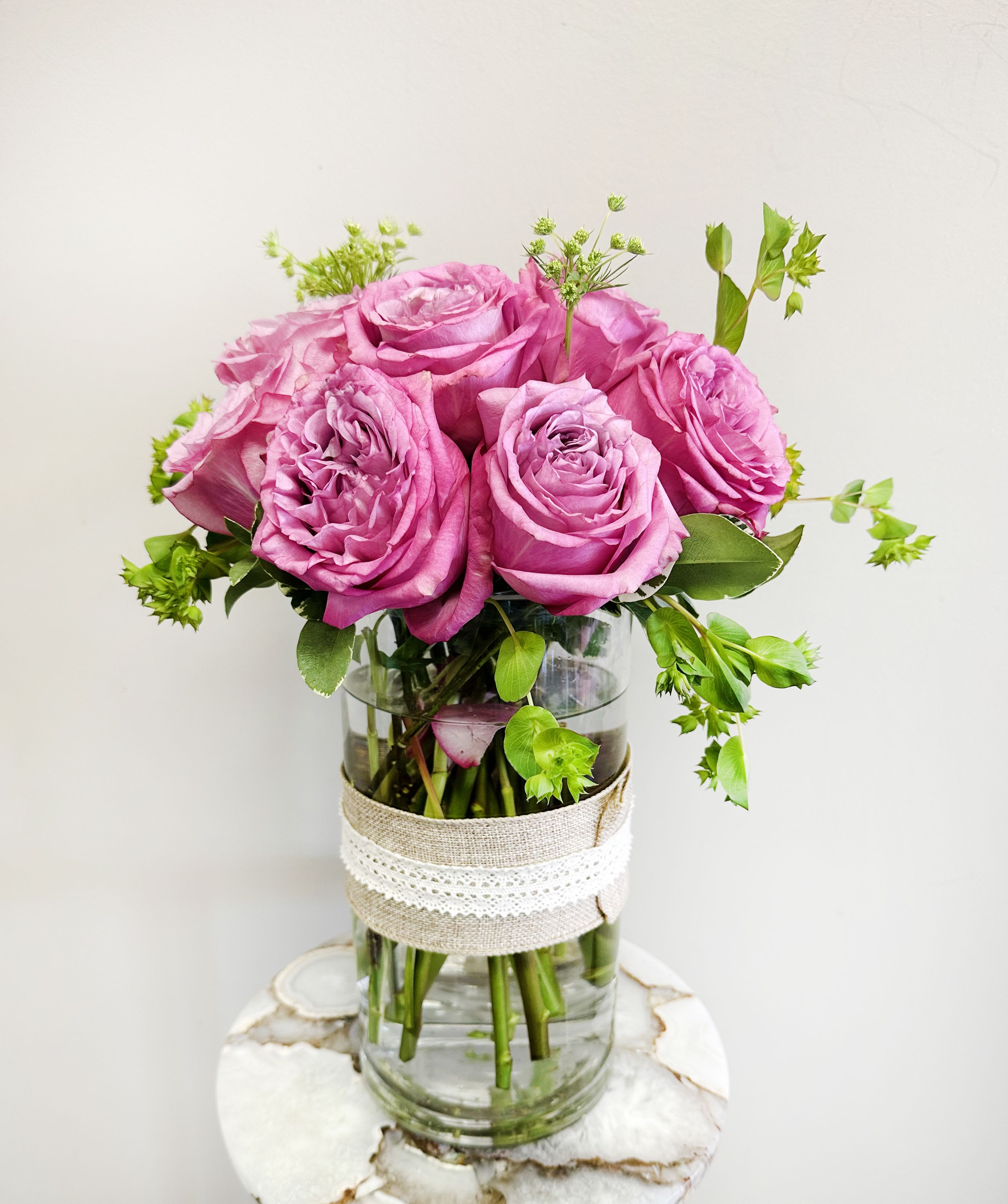 Vintage Lavender Love - Show Mom just how deeply she’s cherished with this graceful bouquet of lavender roses, accented with delicate greens and styled in a tall clear glass vase wrapped in burlap and lace. This arrangement blends timeless beauty with a soft vintage touch—perfect for the mom who’s as elegant as she is down-to-earth. A heartfelt gift that says, “You mean the world to me.” 