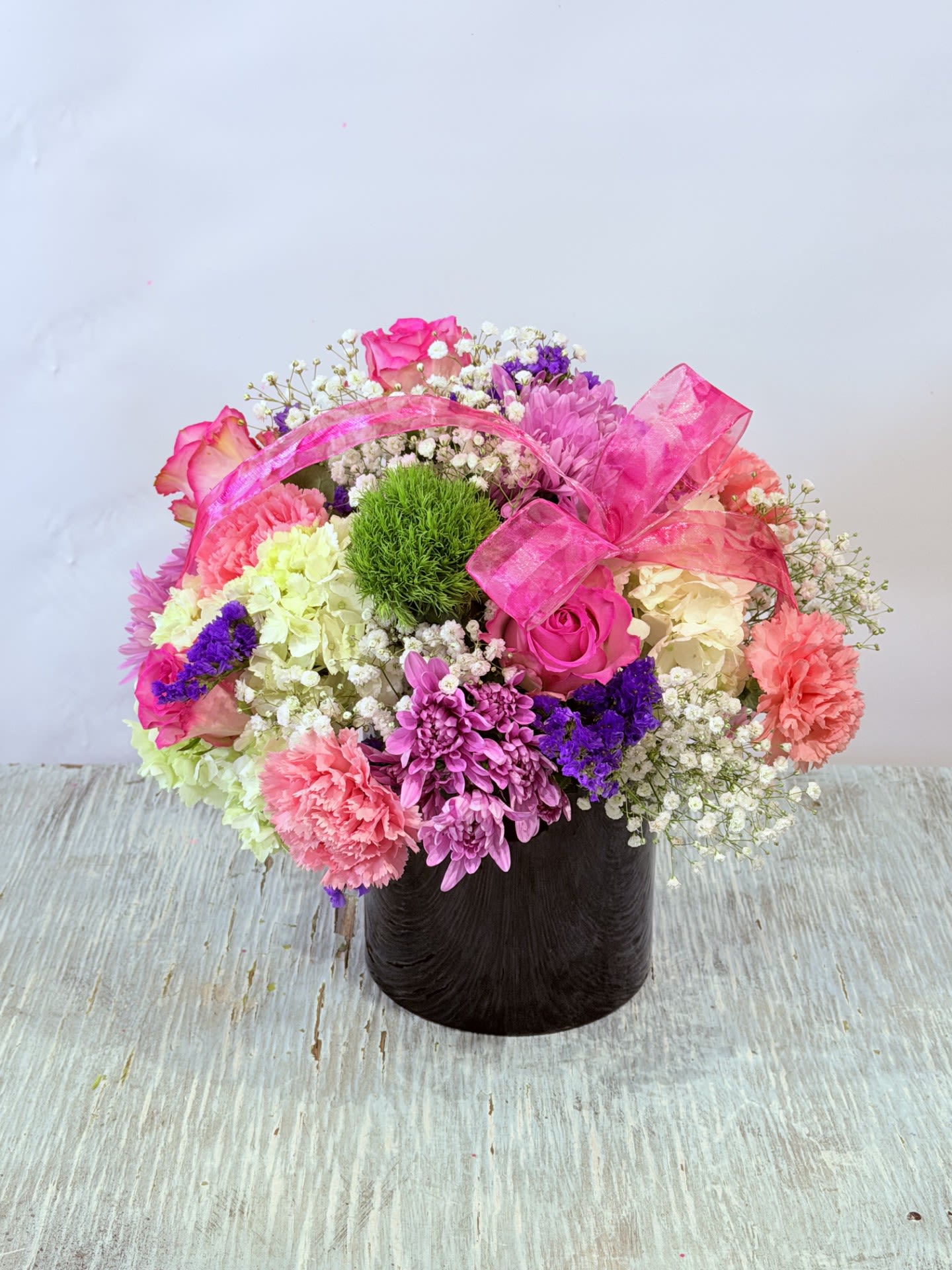 Mother's Day #7 - A cheerful, rounded bouquet of pink, purple, white, and green-this Mother's Day floral arrangement puts soft roses and ruffled carnations front and center for a feminine, celebratory look. Perfect as a Mother's Day gift, birthday bouquet, or thank-you arrangement, it reads as thoughtfully festive and will make the person you're thinking of feel noticed and loved the moment it arrives.  Hand-gathered into a dark, round container, the dense mix includes roses, carnations, chrysanthemums, daisies, clusters of baby's breath, and cream-toned blooms for contrast, with a bright green, moss-like accent at the center that adds unexpected texture. A sheer pink ribbon draped across the top gives a polished, gift-ready finish; the low, cylindrical vase keeps stems neatly arranged and easy to display on a table or mantel.  This arrangement suits moms, grandmothers, friends, or anyone who appreciates soft pastels and a compact, lush presentation-it's the kind of bouquet that makes her feel truly celebrated. Order with standard sizing and expect a long-lasting, easy-to-care-for centerpiece; refresh the water every couple of days and trim stems slightly to keep blooms vibrant.