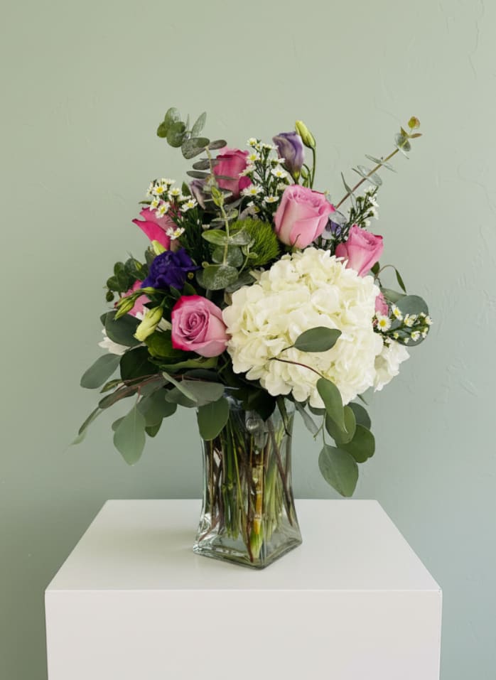 Almond Mom - A full, garden-style bouquet built around a voluminous white hydrangea makes this Mother's Day flower arrangement feel tender and thoughtfully composed. Almond Mom is a mothers day flower arrangement (ideal for Mother's Day, anniversaries, or a thoughtful thank-you) that greets her with soft pink roses, purple lisianthus and roses, and airy white filler, all lifted by eucalyptus for a fresh, fragrant green touch-she'll feel seen and cherished the moment the vase arrives.  Rendered in a clear square glass vase, the design reads both polished and relaxed: a dense white hydrangea focal point anchors the rounded silhouette while some stems reach upward for a loose, garden-picked look. Colors include white, soft pink, purple, and fresh green; flowers are roses, hydrangeas, lisianthus, and delicate fillers. The clear vase shows the stems for an upright, contemporary presentation and makes it easy to display on a table, mantle, or bedside.  Perfect for the mom who prefers romantic, understated elegance, this arrangement sends a message of appreciation and warmth without being over-the-top. Order with confidence for Mother's Day delivery; the arrangement arrives ready to enjoy-just add fresh water and trim stems occasionally to extend vase life.
