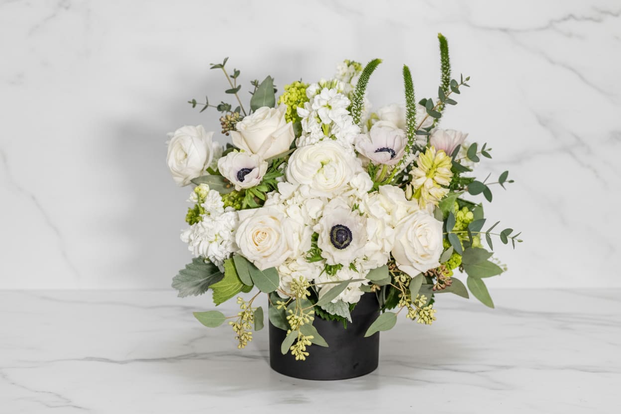 Diamonds and Pearls - “Diamonds and Pearls” is an elegant white floral arrangement featuring lush roses, ranunculus, and anemones accented with soft greenery. Designed in a sleek black vase, this timeless bouquet brings refined luxury to weddings, special occasions, or modern home décor. Perfect for those who adore classic white blooms with a touch of sophistication — available for delivery in San Francisco.