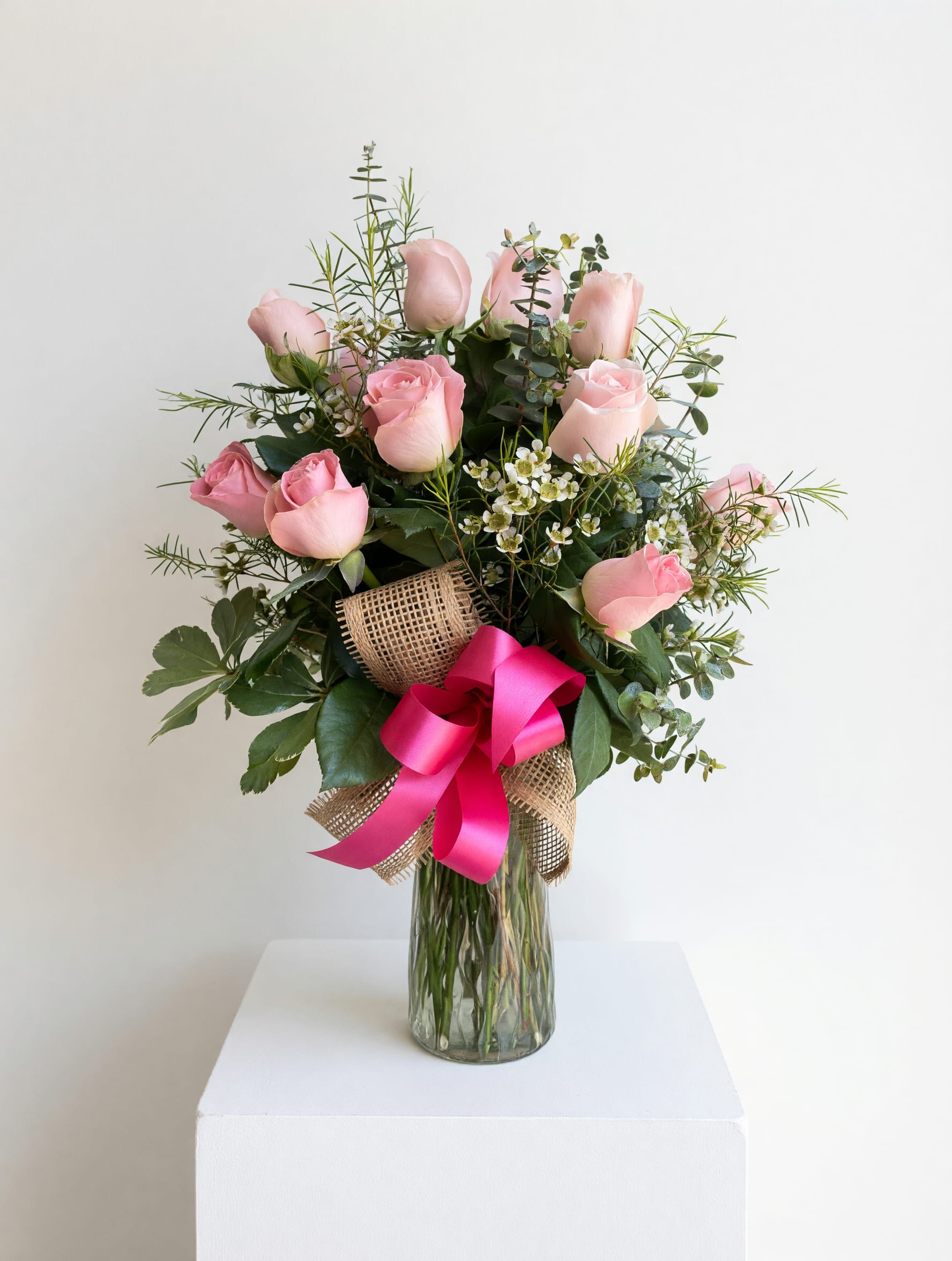 Besitos - Soft pink roses gathered in a loose, airy cluster give this flower arrangement a soft, romantic feel perfect for Mother's Day, birthdays, anniversaries, or a simple "thinking of you." The gentle pink and white palette reads tender and feminine, while the vivid pink satin bow adds a celebratory pop-the kind of bouquet that makes the person you're thinking of feel genuinely cherished.  Clusters of soft pink roses are paired with fine-textured filler and mixed greenery for a garden-fresh silhouette. Stems are arranged in a clear glass vase wrapped in rustic burlap and finished with a bright pink satin ribbon, blending country charm with polished detail. The arrangement's size and airy design create a light, elegant presence on a table, mantle, or bedside.  Made to arrive ready to display, Besitos is an ideal Mother's Day gift that says "thank you" with warmth and sweetness. For longest enjoyment, keep the water fresh and trim stems every few days to preserve that freshly-picked look.