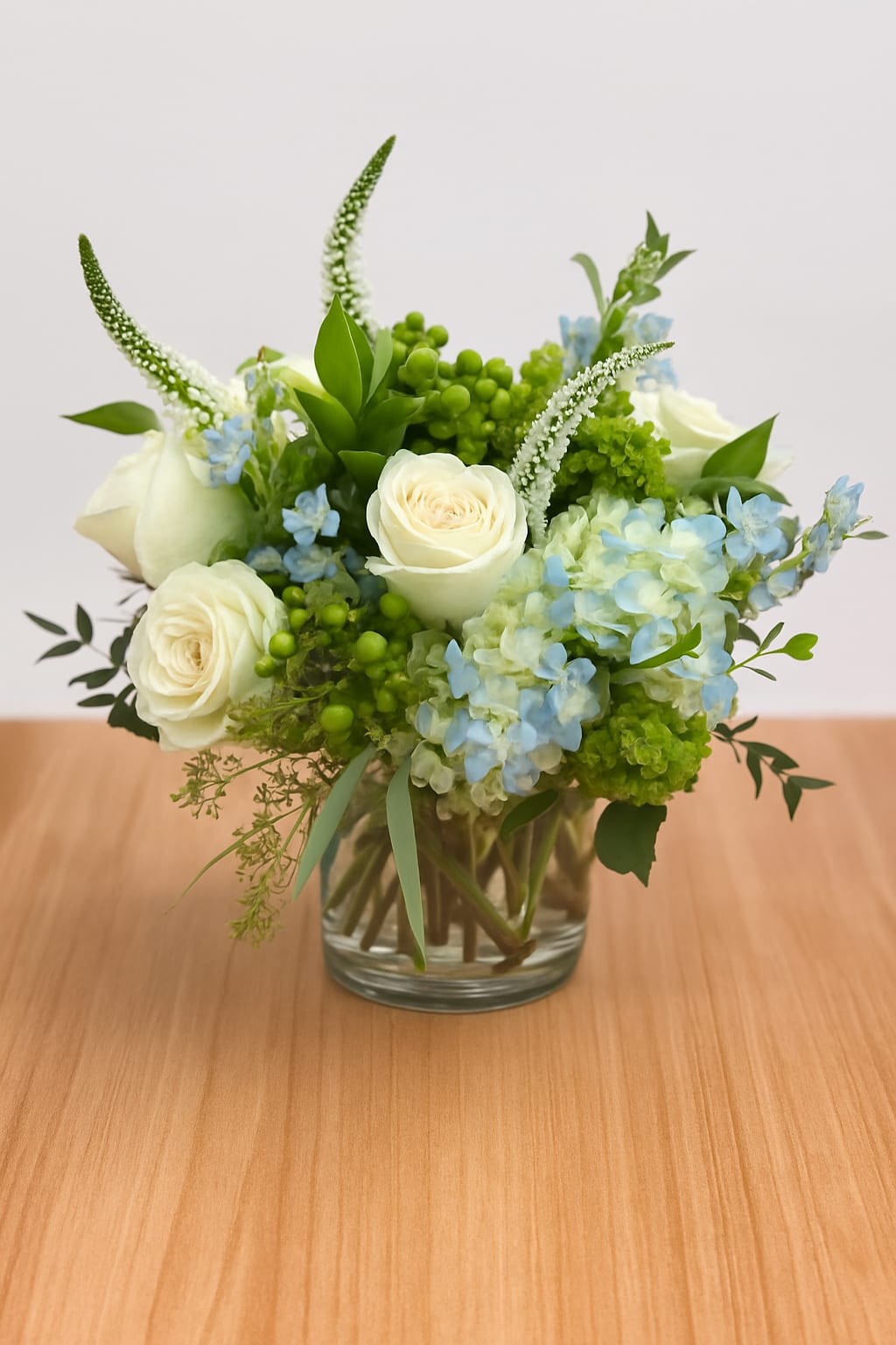 Low round arrangement of white roses, blue hydrangeas, and green accents in a clear glass vase.