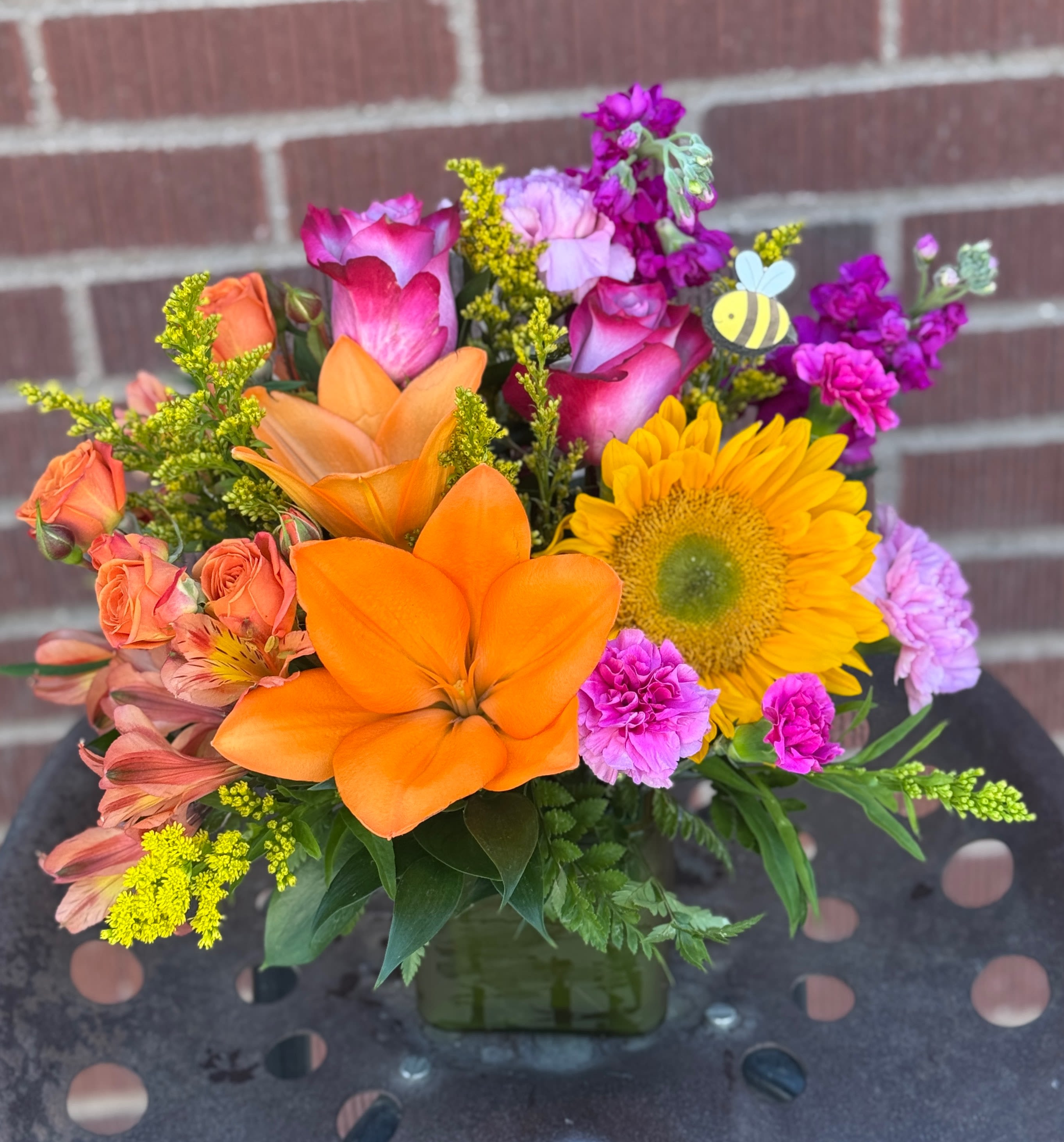 Colorful bouquet with orange lilies, yellow sunflower, and pink roses in a square vase