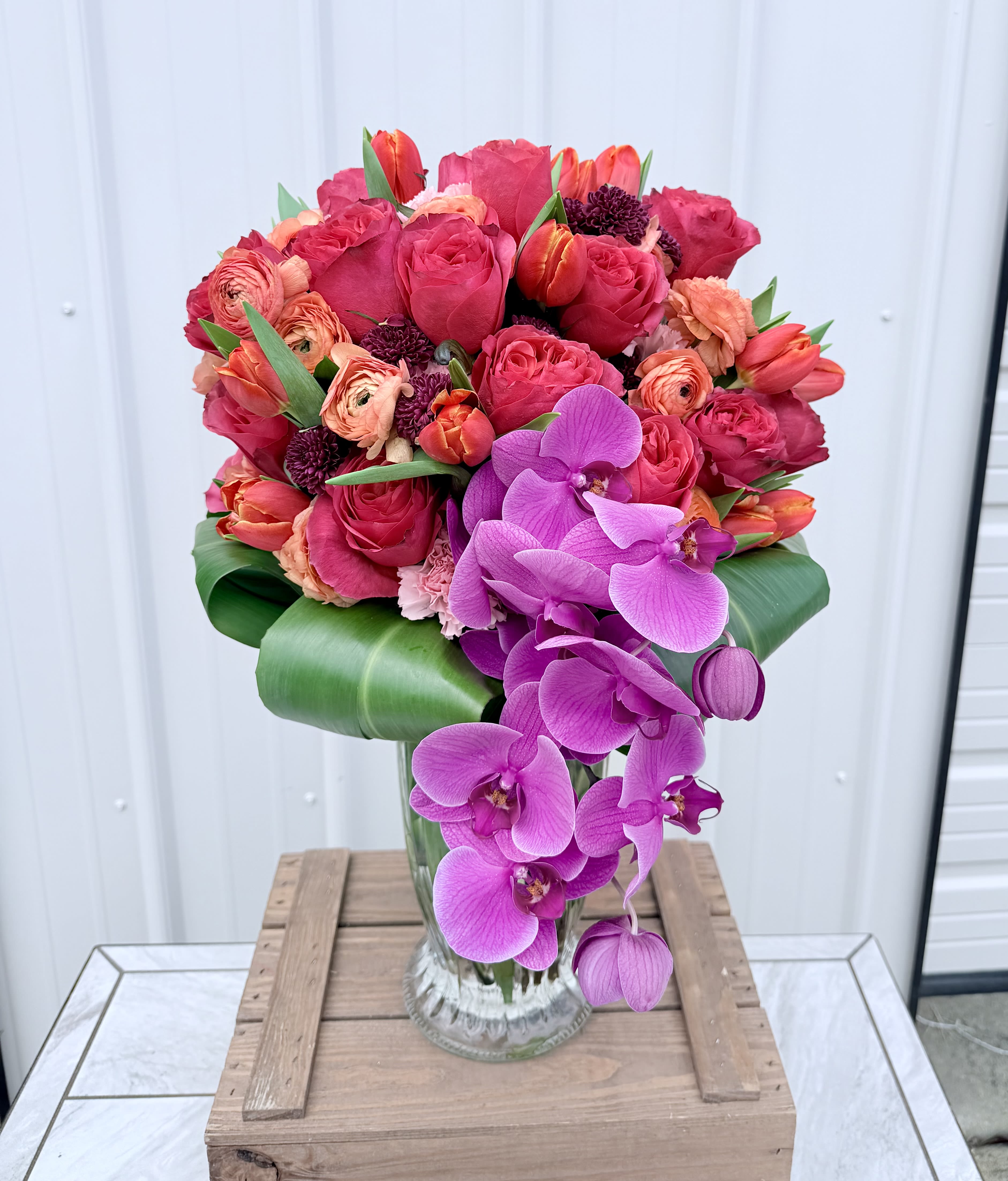 Tropical Radiance - A bold cascade of vivid purple Phalaenopsis and dendrobium orchids drapes over a dense, rounded cluster of hot pink roses, coral ranunculus and orange-pink tulips-this flower arrangement for Mother's Day, anniversaries, birthdays or a "just because" surprise delivers tropical radiance the moment it arrives. The vibrant mix of pink, peach, orange, yellow and white petals reads lively and romantic, so the person you send it to will feel celebrated, seen, and adored whether the occasion is a wedding, graduation, get well wish, or a heartfelt "I'm sorry."  Hand-styled in a clear glass vase, the design pairs tightly layered rose and ranunculus heads with smooth orchid faces for a lush, textured silhouette; broad glossy leaves frame and support the overflowing shape and add a tropical, summer-ready feel. Sized to make a confident statement on a dining table or bedside, this mixed-color arrangement also translates beautifully for sympathy, thank-you gestures, retirement parties, Father's Day with a vibrant twist, and seasonal moments like Spring or Easter-choose delivery to let someone know you're thinking of them.