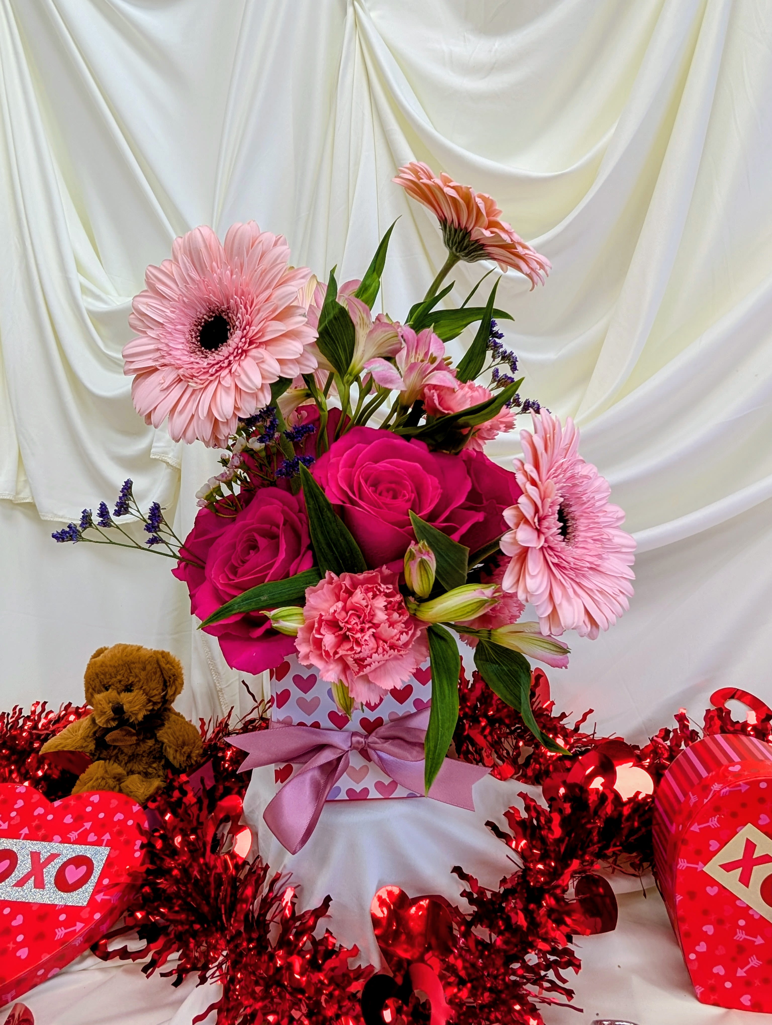 Pink roses and gerbera daisies in a heart-patterned gift box with a teddy bear
