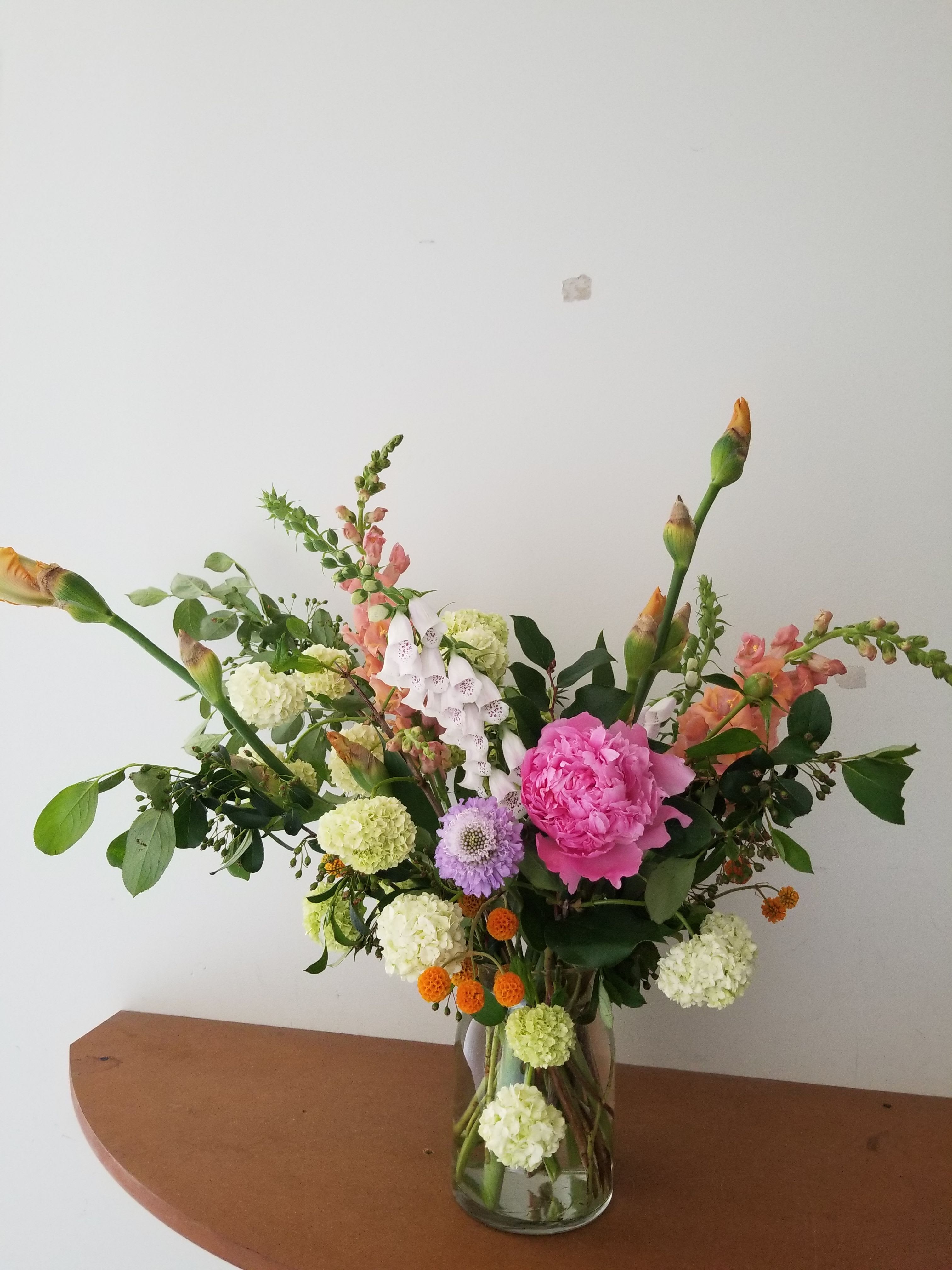 Tall mixed arrangement of pink peony, white hydrangea clusters, snapdragons, and orange accents in a clear glass vase