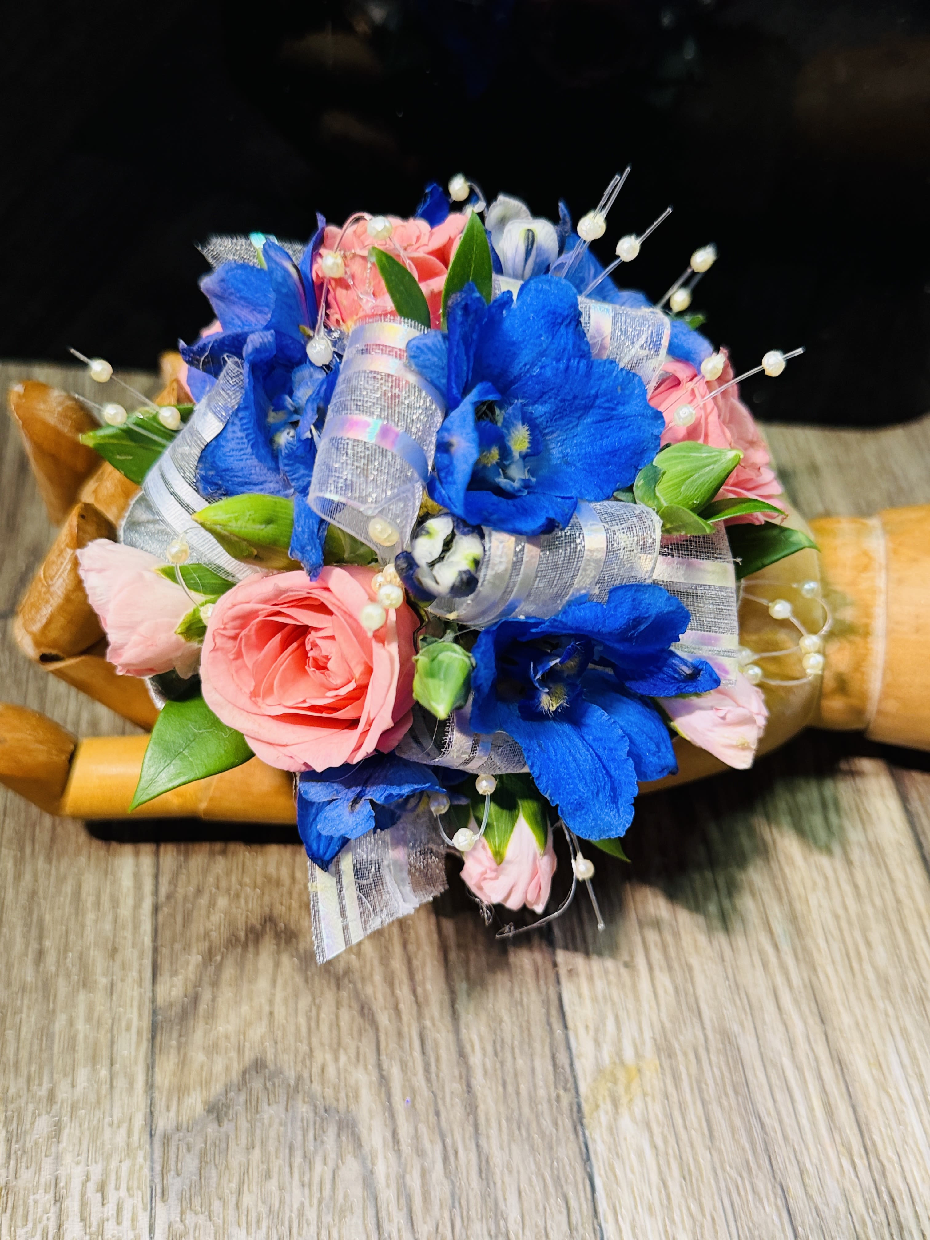 Moonlight Blush Corsage - A delicate and romantic corsage featuring soft light pink roses paired with airy blue delphinium, creating a beautiful balance of warmth and cool tones. The blooms are accented with lustrous pearl details that add a subtle touch of elegance and shimmer. Designed on a graceful pearl wristband, this piece offers both comfort and sophistication, making it perfect for proms, weddings, or any special occasion. The overall look is timeless, feminine, and effortlessly charming—like a miniature garden wrapped around your wrist.