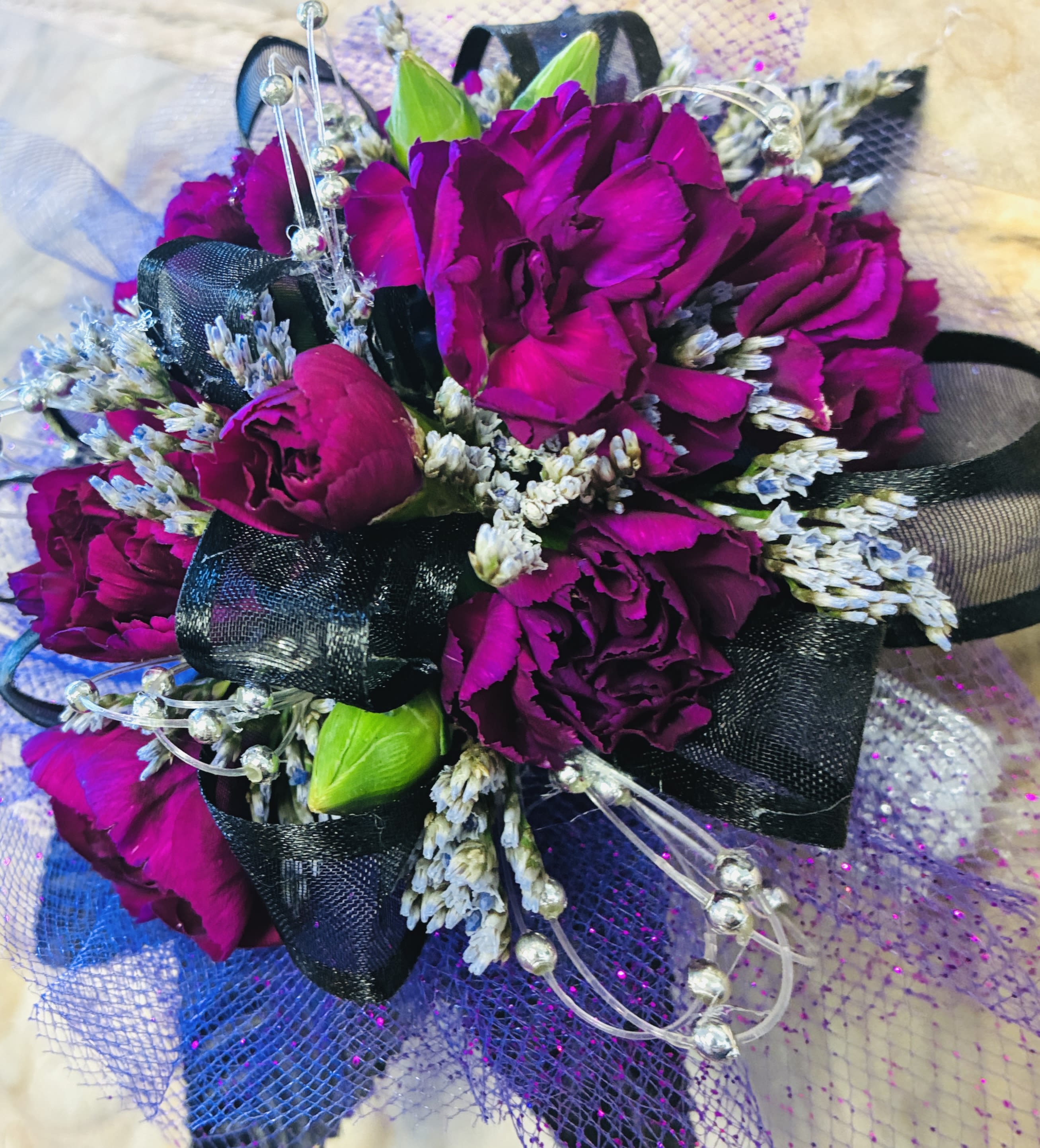 Viper Elegance  - The carnation itself is the focal point—its tightly layered petals in a deep plum or almost-black purple create a velvety texture that catches light subtly, giving it depth rather than brightness. The color leans toward moody and luxurious, reminiscent of twilight or crushed velvet. Wrapping the base, the black ribbon adds contrast and structure. It can be satin for a sleek shine or matte for a softer, gothic feel. The ribbon may be tied into a neat bow or wrapped cleanly around the stem, grounding the design with a bold, dark frame. Threaded through or clustered near the bloom, silver beads bring a delicate sparkle. They might appear as tiny accents nestled between petals or strung on thin wires that arc gently outward, catching the light like small stars. This metallic touch breaks up the darkness just enough, adding refinement and a hint of glamour. Overall, the corsage feels sophisticated, slightly gothic, and evening-ready—perfect for formal events where you want something understated but striking.