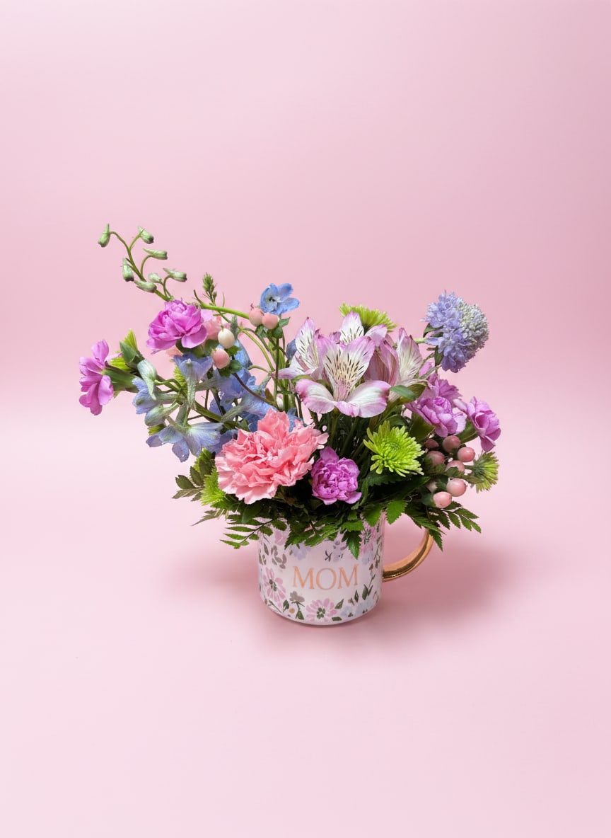 Mom’s Meadow - A soft and uplifting Mother’s Day mug arrangement designed to feel like a gentle stroll through a blooming spring garden. Presented in a keepsake coffee mug featuring delicate pink and blue floral artwork with the word “Mom,” this piece blends pastel color with natural, garden-inspired texture.  Lavender mini carnations and lavender scabiosa bring a light, airy calmness, while pink alstroemeria and pink carnations add warmth and a cheerful blush of color. Green cushion/Kermit mums provide fresh texture and contrast, and peach hypericum berries offer a soft, playful accent that ties the palette together beautifully.  Thoughtful, fresh, and full of heart, this arrangement is a sweet little meadow moment made just for Mom—finished in a reusable mug she’ll enjoy long after the flowers are gone.  All flowers, flower colors, and containers are subject to change due to availability.