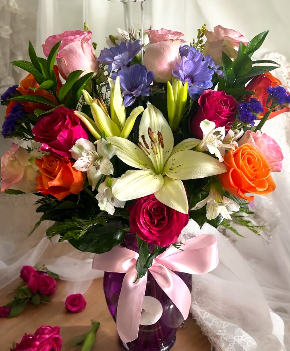 Mixed bouquet of roses and lilies in a purple vase with a pink ribbon