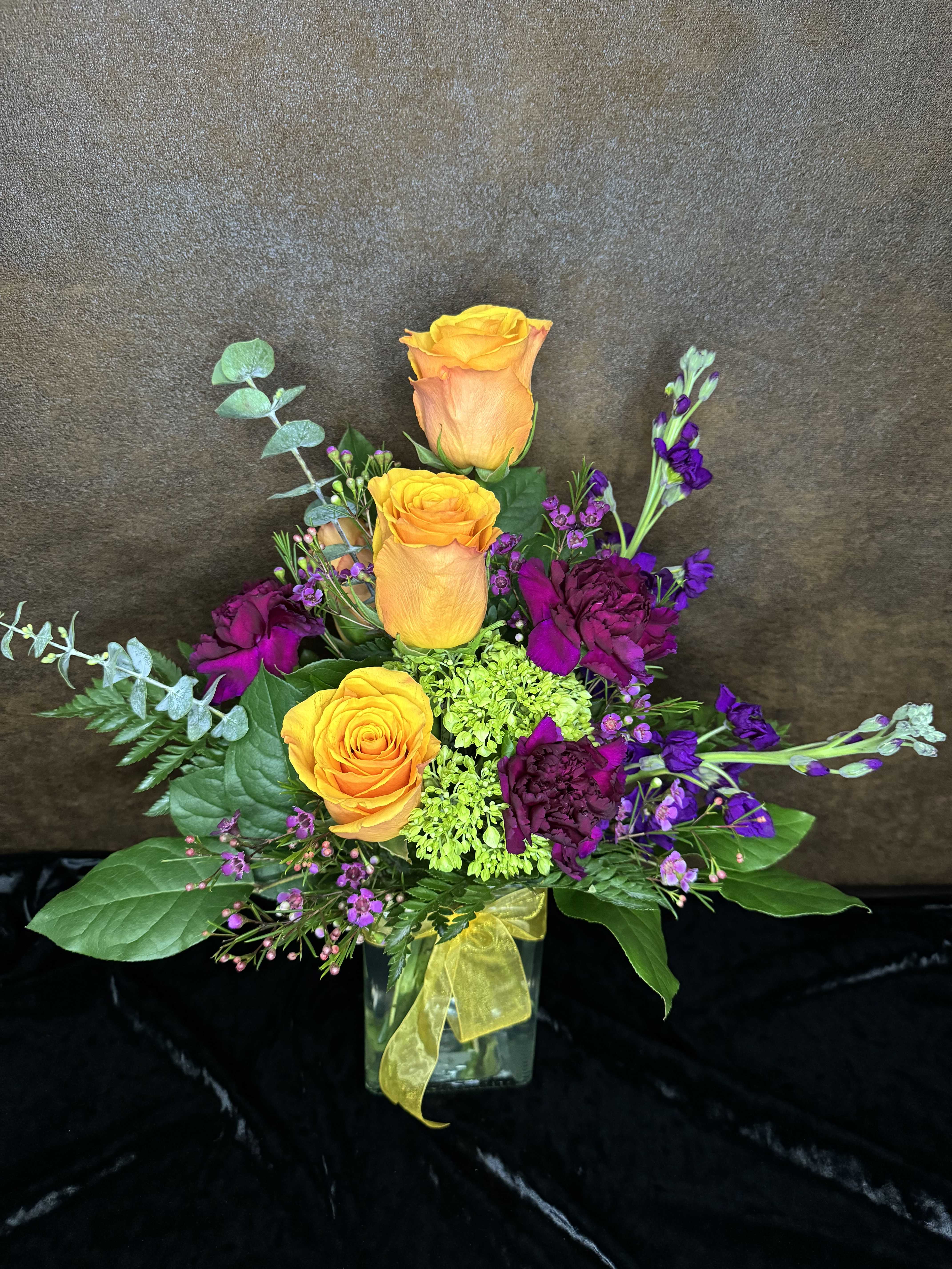 Orange roses and purple flowers in a glass vase with a yellow ribbon