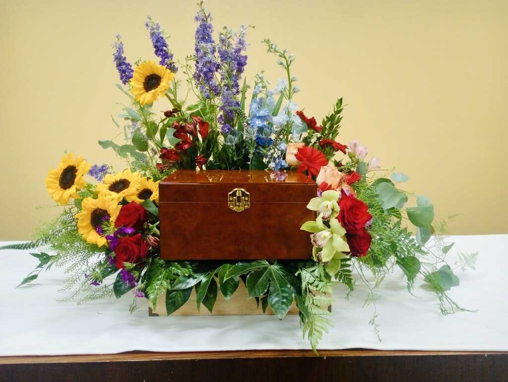 Celebration of Life Box - A vibrant, living tribute centered around a polished wooden keepsake box, this flower arrangement is a Celebration of Life box perfect for sympathy and funeral services or a bright memorial display for Memorial Day. The asymmetrical, garden-like composition uses tall purple and blue delphinium spikes and rich red roses to give height and dignity, while golden sunflowers and mixed-color alstroemeria bring warmth and lift-creating the kind of bouquet that makes the person you're honoring feel vividly remembered.  Surrounding the wooden Urn box  the design layers pale orchids, sunflowers, roses, delphinium, and complementary greenery for a lush, expansive silhouette that softens the box's lines. The piece reads both formal and celebratory: dense, trailing foliage frames the arrangement so it sits beautifully on a memorial table, beside a casket, or at a home altar for Dia de los Muertos or Día de las Flores Amarillas. Sized to make a statement without overwhelming a space, it arrives ready to display and keeps the keepsake box at the heart of the tribute, offering a colorful, comforting way to say you're thinking of them.
