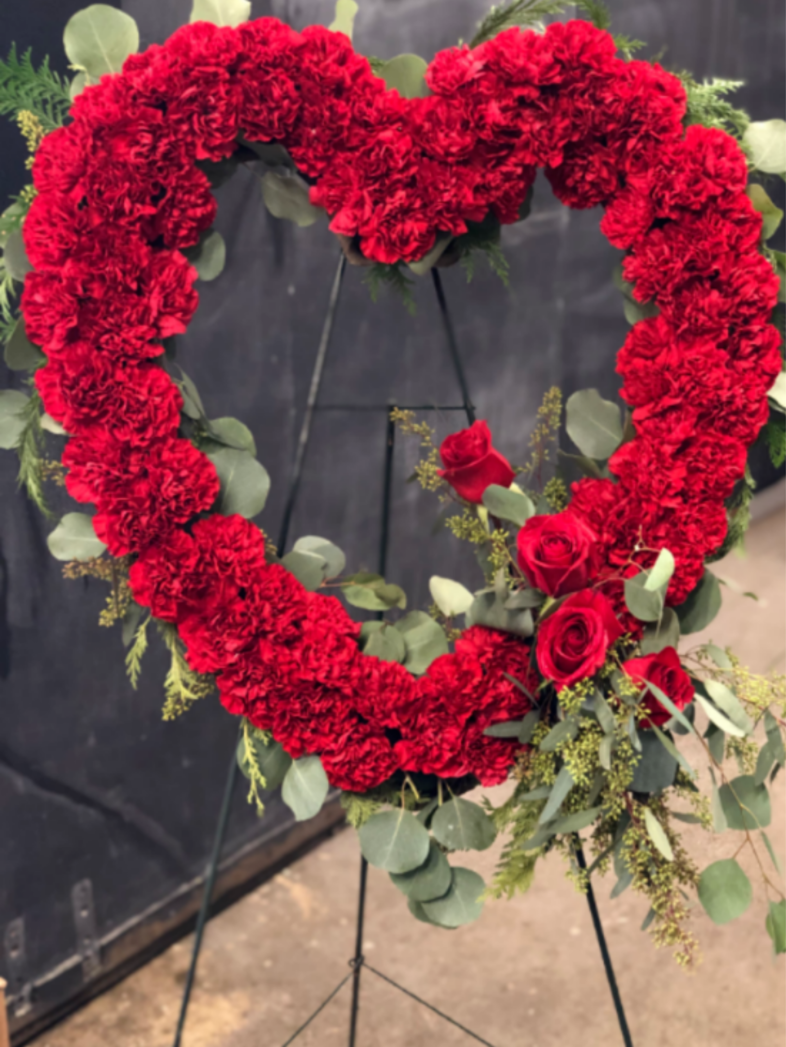 Sympathy Heart - A bold heart of densely packed red carnations-accented with a cluster of red roses-creates an unmistakable sympathy arrangement and funeral tribute that reads as both romantic and reverent. The heart shape and vivid crimson color immediately convey love and remembrance, making this flower arrangement a meaningful choice for a memorial service, a Funeral program, or a Memorial Day tribute to honor someone special.  Soft eucalyptus-style foliage and airy filler greens frame the silhouette, adding muted green contrast and gentle texture against the vivid red blooms. The piece is mounted on a sturdy black metal easel for formal presentation and easy display at a service or graveside. Designed to feel dramatic without being ornate, it's the kind of tribute that will make the person you're remembering feel deeply honored and seen.  Order sizing and delivery options are available at checkout; the metal easel is included so the arrangement arrives ready to display. For best appearance at the service, keep the arrangement in a cool, shaded spot before placement-its densely packed carnations and roses are chosen for lasting color and presence during memorial events.