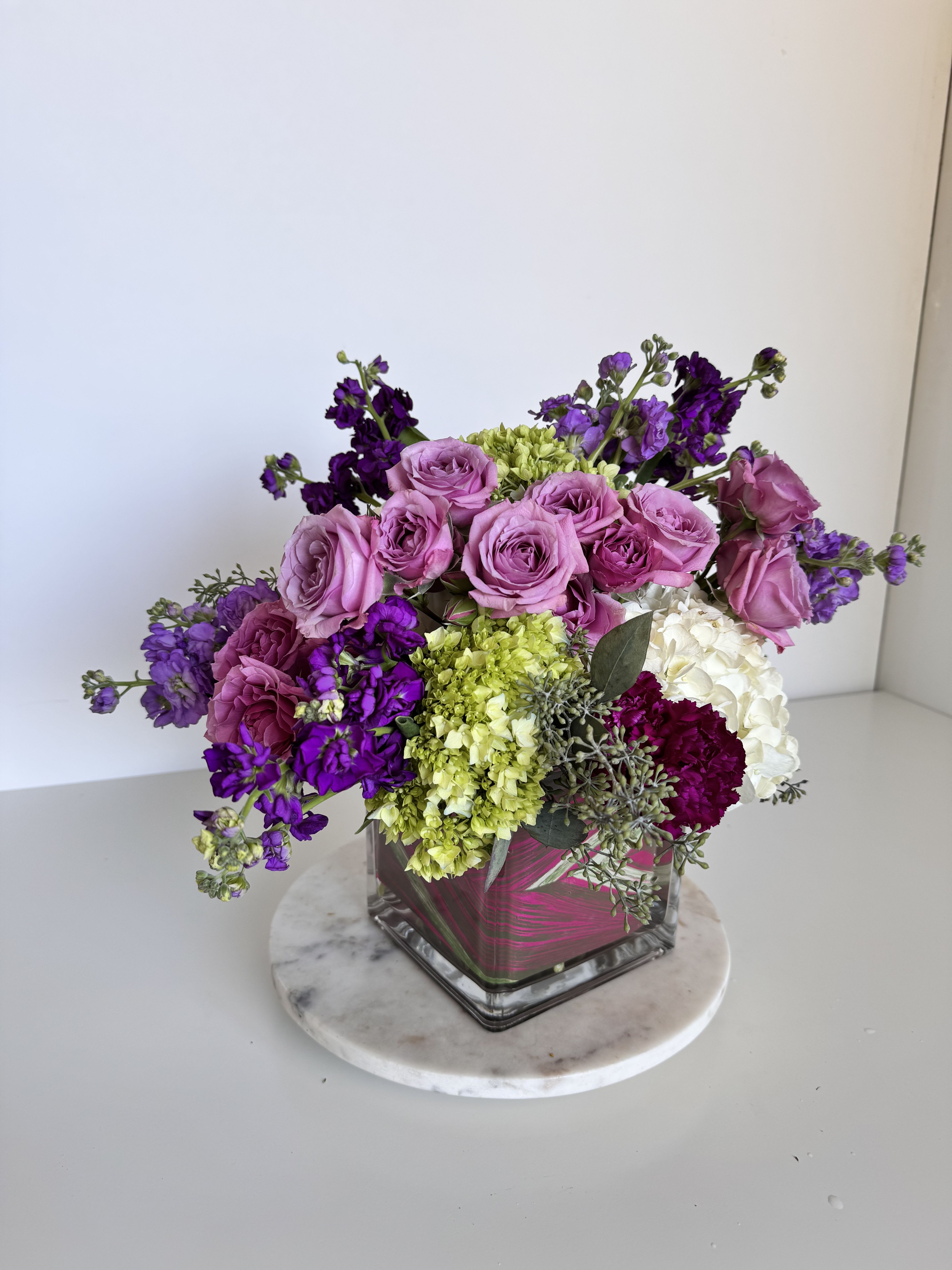 Purple Delight - A lush cluster of blush and lavender roses paired with vivid purple stock and pale green hydrangeas makes this birthday bouquet or anniversary arrangement immediately feel celebratory and romantic. Purple Delight is designed to make the person you're thinking of feel cherished-its dense, garden-style composition and pops of white and deep magenta create a joyful, confident statement perfect for birthdays, anniversaries, or a heartfelt thank-you.  This compact arrangement sits in a square glass vase atop a round marble pedestal, giving the full, rounded silhouette a clean, modern base. Layers of pink and purple roses blend with fluffy white and green hydrangea heads, fragrant stock, and textural carnations to create depth and movement-some stems arch gently outward for a natural, freshly-gathered look. The palette of pink, purple, white, and green reads bright and feminine while still feeling sophisticated.  Choose this bouquet when you want her to feel truly seen-whether celebrating a milestone or sending warm thanks. The size and dense arrangement make it a standout centerpiece on a dining table or a lovely bedside accent; arrange on a stable surface and refresh water every 2-3 days for best vase life.