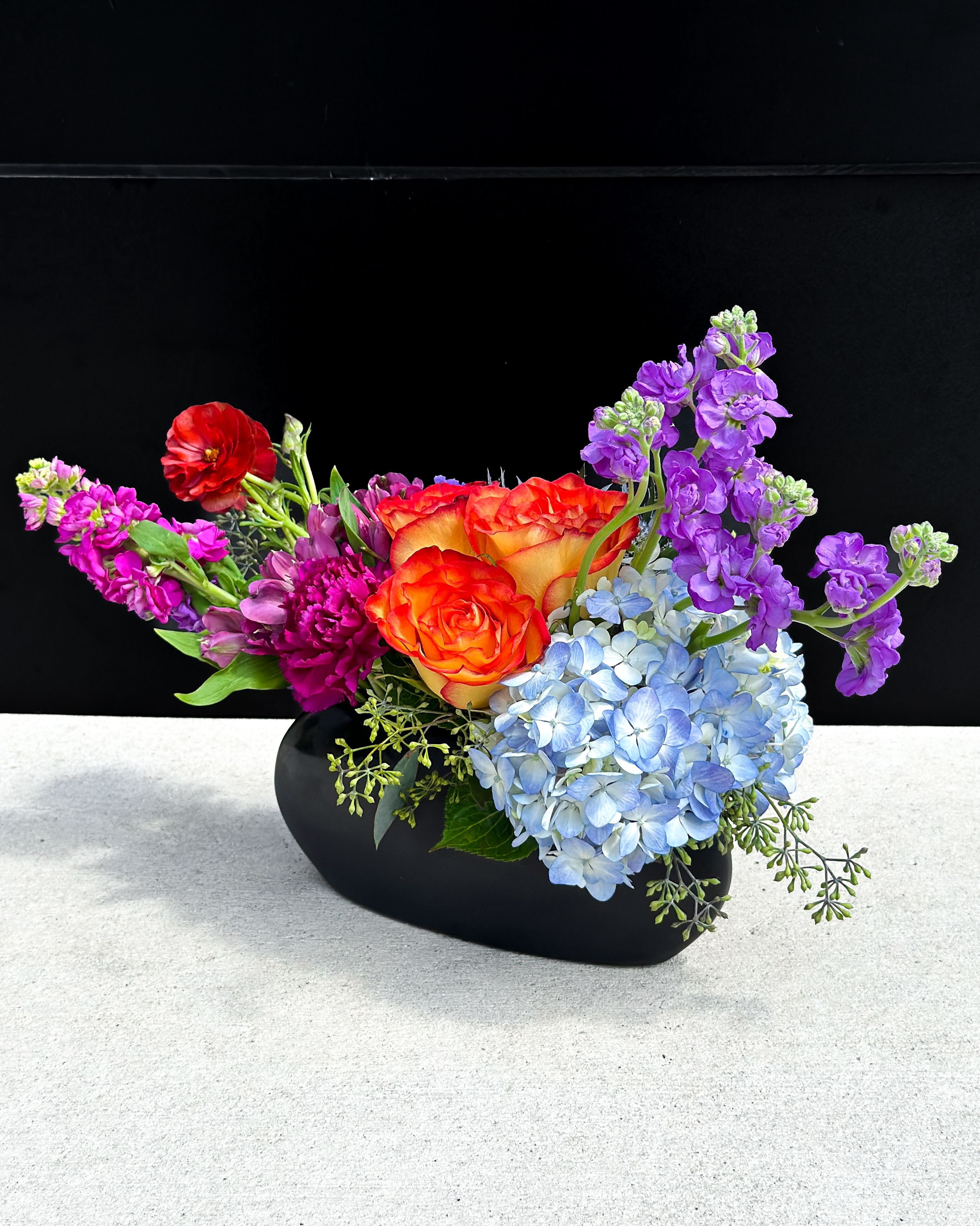 Sunset Delight - Vivid orange-red roses meet pale blue hydrangea and tall purple stock in a lively mixed bouquet built to brighten birthdays, congratulations, or any celebratory moment. The Sunset Delight birthday bouquet reads like a joyful, modern painting-bold color contrasts and an asymmetrical, abundant silhouette that makes the recipient feel genuinely celebrated the moment they see it.  This arrangement pairs soft-textured hydrangea blooms in pale blue with velvety orange-red roses, upright purple stock, and smaller magenta and red accents for depth and movement. Displayed in a sleek black vase, the composition gains a contemporary, dramatic anchor that highlights the colors and keeps the overall look sophisticated yet playful. Approximate dimensions and care information are available at checkout; stems arrive arranged and ready to display, with simple water care to keep the flowers fresh for as long as possible.