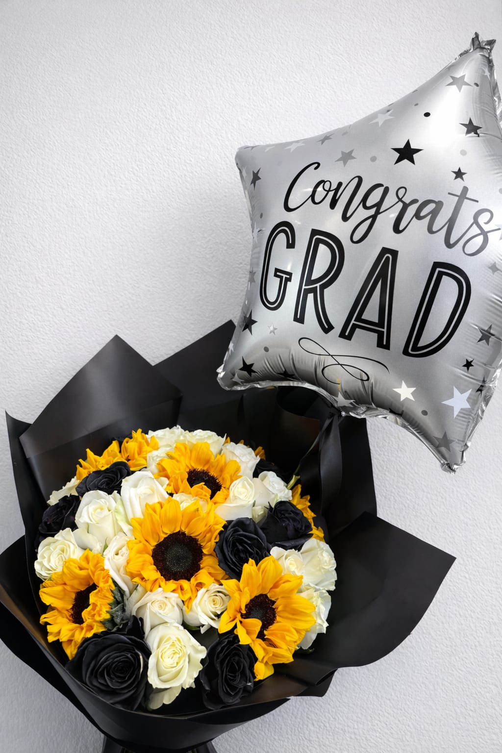 Graduation Celebration Bouquet  - Celebrate their big achievement in style with this bold and elegant Graduation Bouquet. Featuring a stunning mix of vibrant sunflowers, classic white roses, and striking black roses, this arrangement is designed to make a statement and honor their special milestone.  Wrapped in sleek black paper for a modern, sophisticated look, this bouquet perfectly balances brightness and elegance—symbolizing success, new beginnings, and a future full of possibilities. Paired with a festive “Congrats Grad” balloon( Balloon Color May Vary depending on Availability), it’s the perfect all-in-one gift to surprise and celebrate your graduate.   Perfect for:  Graduation ceremonies Senior photos Surprise gifts Celebration parties  Make their moment unforgettable with a bouquet as bright and bold as their future   Contains : 24 Roses and 6 Sun Flowers 