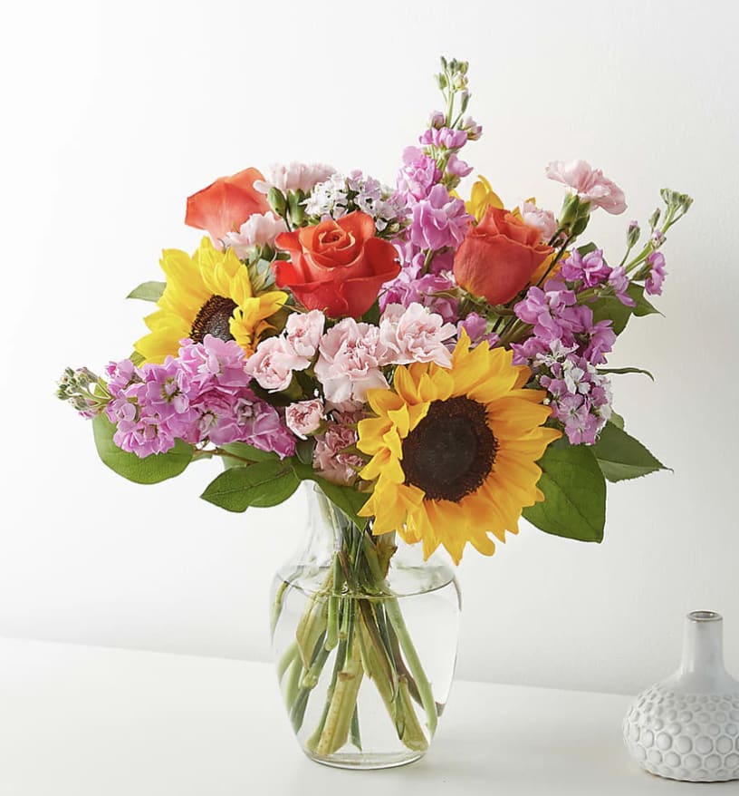 Mixed bouquet of sunflowers, orange roses, and pink blooms arranged in a clear glass vase.
