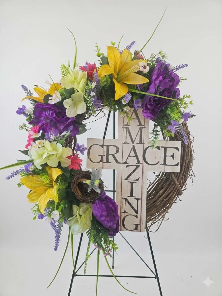 Amazing Grace Grapevine Wreath  - A reverent, uplifting wreath built around a wooden cross sign that reads "Amazing Grace"-perfect for funeral services, a memorial display, or as inspirational home decor. The artificial flower arrangement wreath pairs bright yellow lilies with rich purple blooms and pale green hydrangea-like clusters, accented by small white filler flowers to create a peaceful, hopeful palette that reads both comforting and celebratory for the person you're honoring.  Set on a rustic grapevine wreath, the design is full and layered with long stems and gentle trailing accents for a handcrafted, natural feel. The wooden cross sign is the focal point, and the wreath ships ready to display on the included metal easel for easy placement at a service or on a mantel. Sized to make a meaningful statement without overwhelming a space, this arrangement gives the recipient-or the memory you're honoring-a graceful, beautifully composed tribute. 22" Around Wreath