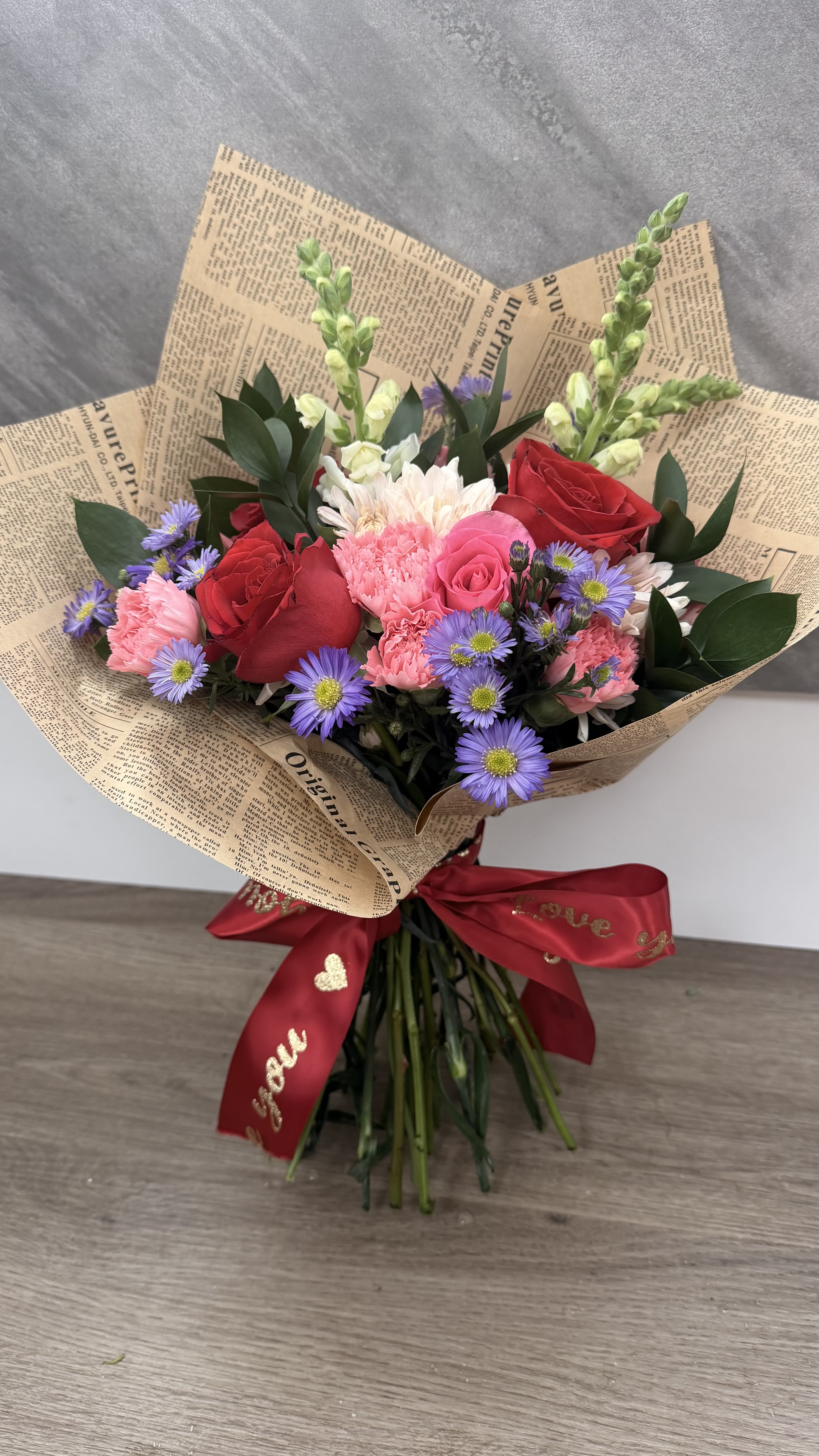 Hand-tied bouquet of red roses, pink carnations, and purple asters wrapped in printed paper with a red ribbon