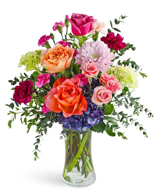 Harmony - A lively mix of pink, orange, purple, and white blooms creates an immediate lift-perfect as a birthday bouquet, a cheerful thank-you, or a "just because" surprise. The flower arrangement features garden-style abundance and a bright, layered palette that makes the person you're thinking of feel seen and celebrated.  Large roses anchor the design while ruffled carnations and a full hydrangea cluster add volume and softness; a pale chrysanthemum brings delicate contrast and texture. Airy greenery extends outward for a loose, natural silhouette, all arranged in a clear glass vase that showcases the stems and complements any room.  Sized to make an impression without overwhelming a tabletop, this arrangement reads as both joyful and thoughtfully composed-the kind of gift that brightens a day and invites guests to smile. Simple care: fresh water in the vase and a weekly trim will keep the blooms looking their best.