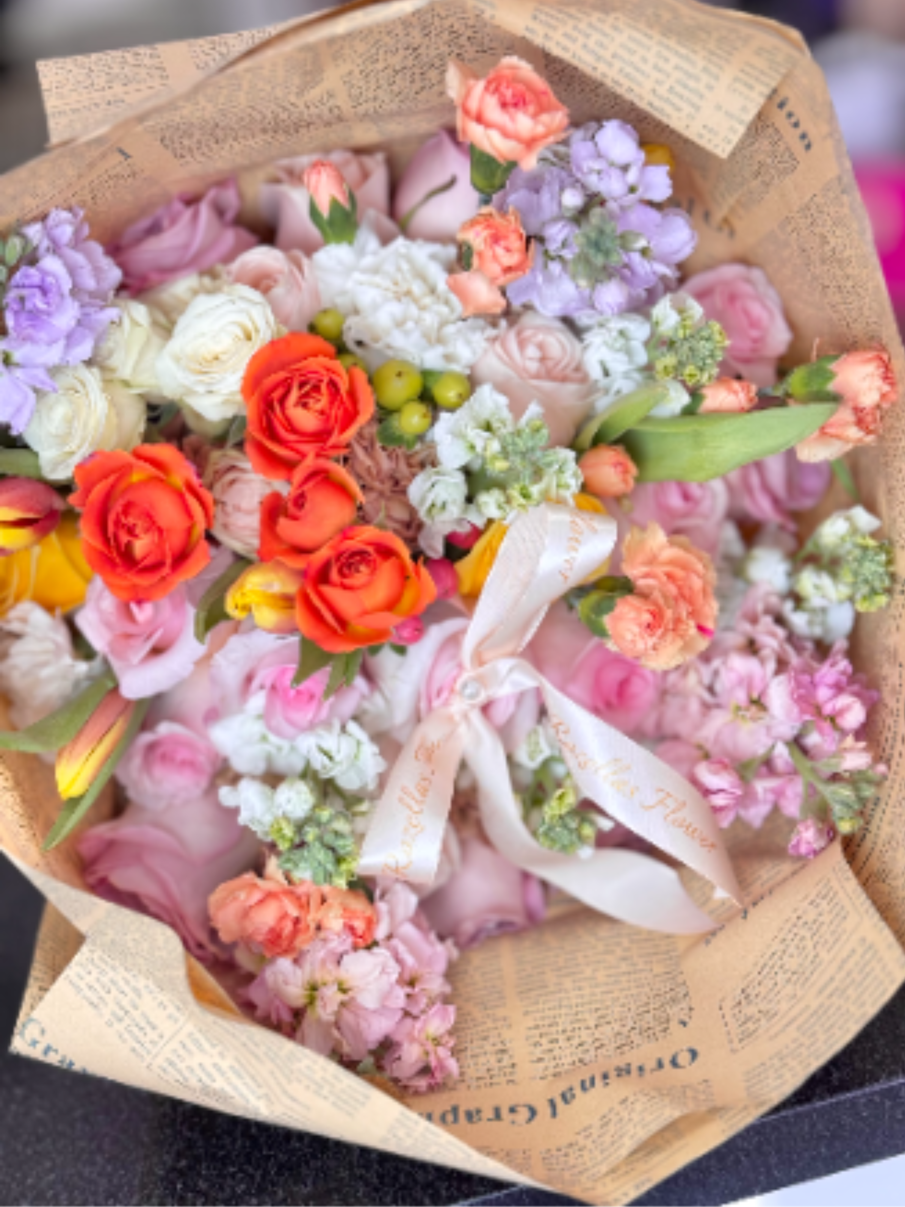 Blush Bouquet - A dense, hand-tied flower arrangement bursting with soft pastels and a pop of bright orange to catch the eye - perfect as a birthday bouquet, anniversary gift, or a special Mother's Day surprise. The rounded, abundant composition reads romantic and joyful, the kind of arrangement that makes the person you're thinking of feel genuinely celebrated and seen.  Roses, carnations, stock and small clustered filler blooms are arranged in pink, white, orange and lavender tones with a few green berry-like accents for contrast. Wrapped in tan printed paper and finished with a pale satin ribbon, the bouquet arrives gift-ready with a plush, tactile presentation that feels thoughtful and polished.  Ideal for springtime gifting, a get-well cheer, or to mark Women's Day or a meaningful milestone, this arrangement suits anyone who loves soft color palettes with a lively twist. Hand-tied for fullness and wrapped to stay neat in transit; treat it gently and refresh the water promptly when you place it in a vase to extend the life of the blooms.
