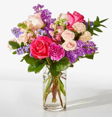 The Cha-Cha Bouquet - A lush, garden-like bouquet of pink, purple, white, and green that reads both cheerful and romantic - perfect as a birthday bouquet, anniversary arrangement, or a just‑because gift to make someone feel truly seen. Large pink and blush roses anchor the design so the recipient is met with classic romance, while pops of bright magenta carnations and soft purple stock add energetic color and fragrant, layered texture.  The Cha-Cha Bouquet is gathered into a clear cylindrical glass vase that shows the stems through the water for a fresh, timeless presentation. Alongside the roses you'll find clustered filler blooms and airy stock that create a full, rounded silhouette with blooms layered at varied heights; the palette mixes vivid pinks, lavender, cream, and leafy green for a polished everyday look. The arrangement feels abundant without being fussy, offering a tactile mix of velvety petals and delicate sprays.  For birthdays, anniversaries, or a thoughtful thank-you, this bouquet gives the recipient an immediate lift - bright, romantic, and unmistakably celebratory. It arrives ready to display in the included glass vase; keep blooms in fresh water, out of direct sunlight, and trim stems slightly every few days to extend vase life.