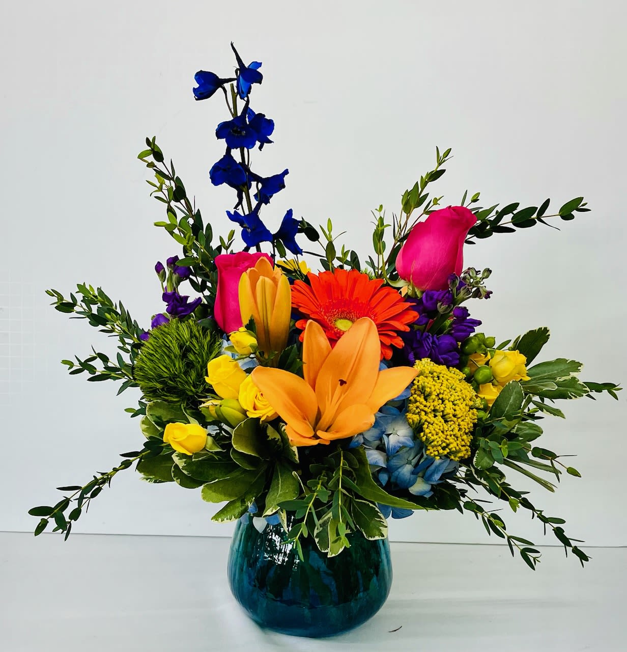 Happy Blooms - Brighten any occasion with our Happy Blooms arrangement—a joyful mix of vibrant orange, purple, blue, pink, and yellow flowers designed to make a lasting impression. This stunning bouquet features a fresh selection of gerbera daisies, roses, lilies, spray roses, hydrangea, hypericum, delphinium, and lush greenery, artfully arranged to create a colorful and uplifting display.  Perfect for birthdays, celebrations, or simply bringing a smile to someone’s day, Happy Blooms delivers beauty and cheer in every stem.  We proudly offer delivery throughout Smyrna, Marietta, Mableton, Austell, and Atlanta, ensuring your flowers arrive fresh and on time.  Please note: Flower substitutions may be made based on seasonal availability while maintaining the overall look, feel, and color palette of the arrangement.