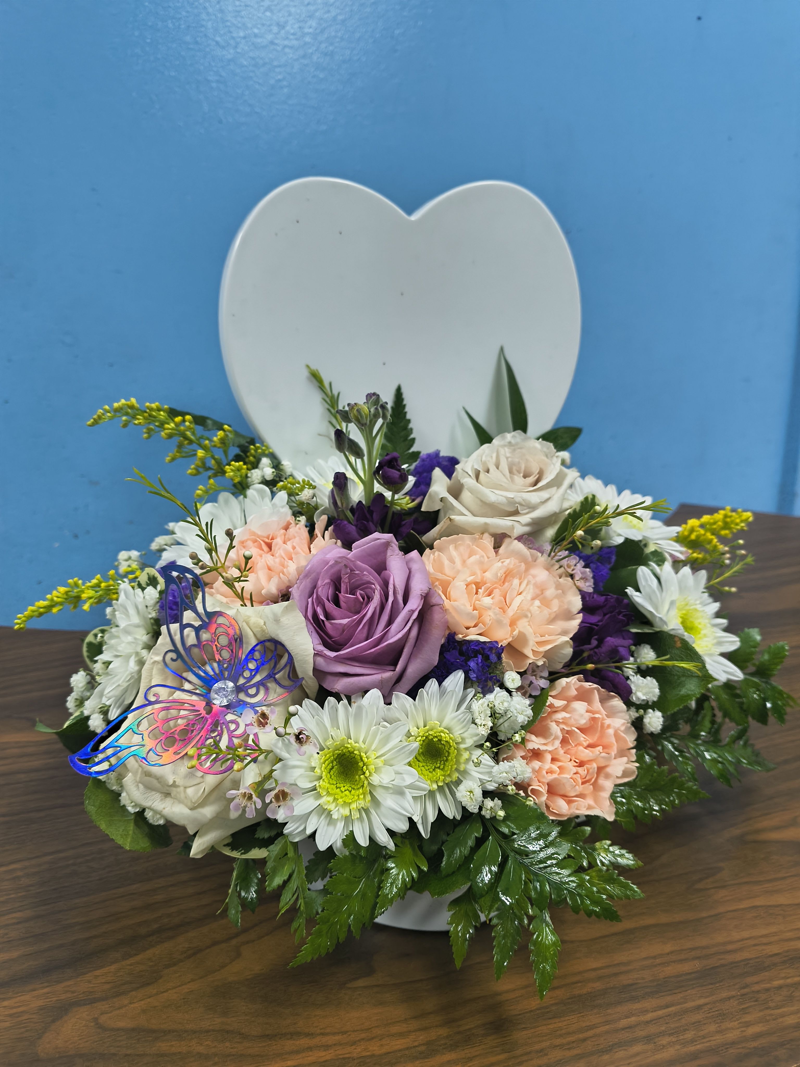 A Mothers Love - Creamy roses and soft lavender blooms nestle against a white heart-shaped plaque in this compact Mother's Day flower arrangement, an ideal gift to make the person you're thinking of feel cherished and seen. The bouquet reads like a warm embrace - peach and white petals soften the palette while bright yellow accents lift the mood, so it works just as beautifully for an anniversary, a thank-you, or any sentimental moment.  This low, wide design layers roses, carnations, and daisy-like flowers to create a textured, romantic look with playful pops of color. A delicate decorative butterfly pick adds a whimsical touch to the heart backdrop, making the arrangement feel personal and keepsake-worthy. Perfect for tabletops or a bedside table, it arrives ready to display and brings a fresh, fragrant presence to the room.