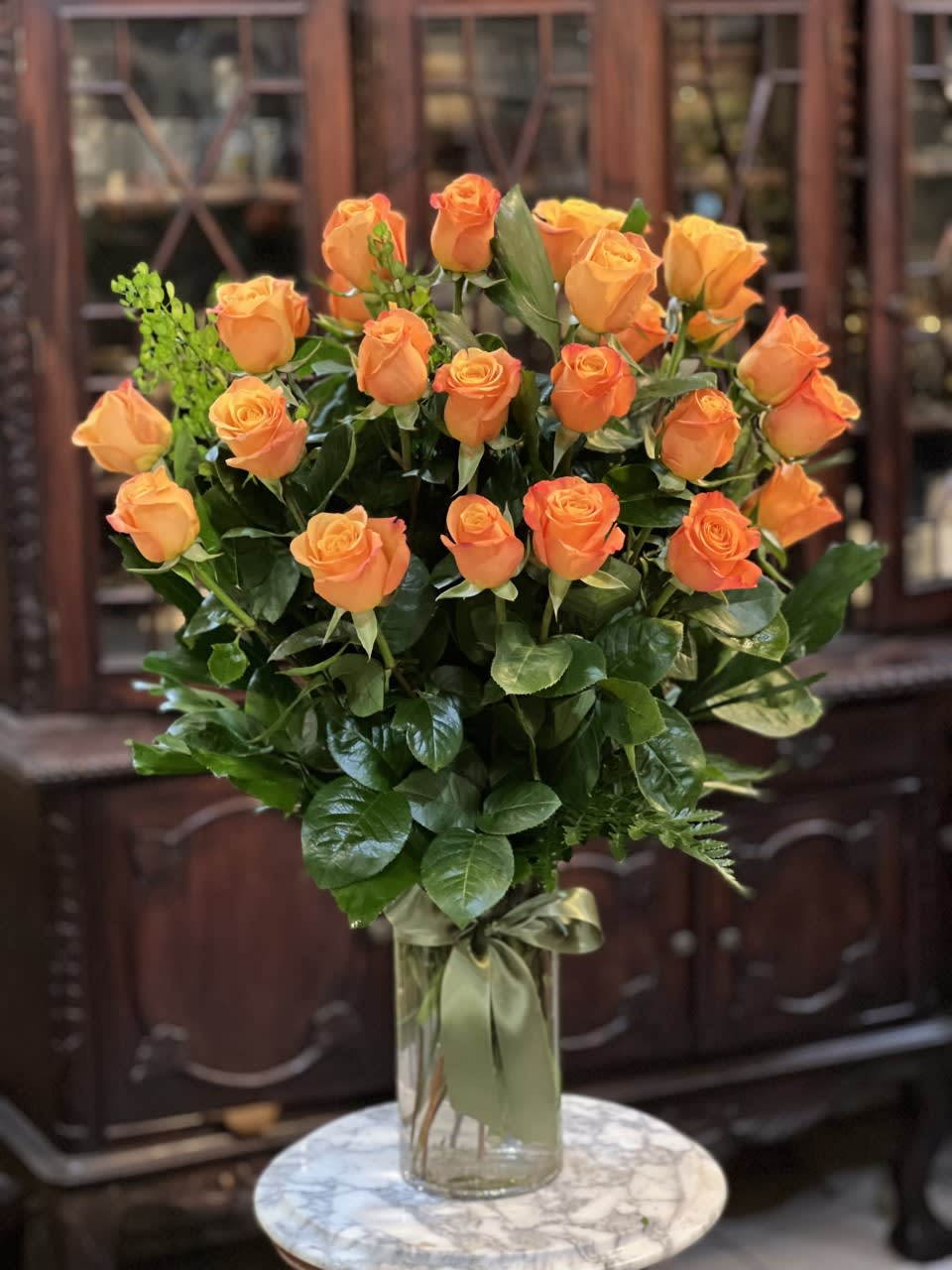 Tall arrangement of orange roses in a clear glass vase with a green ribbon bow.