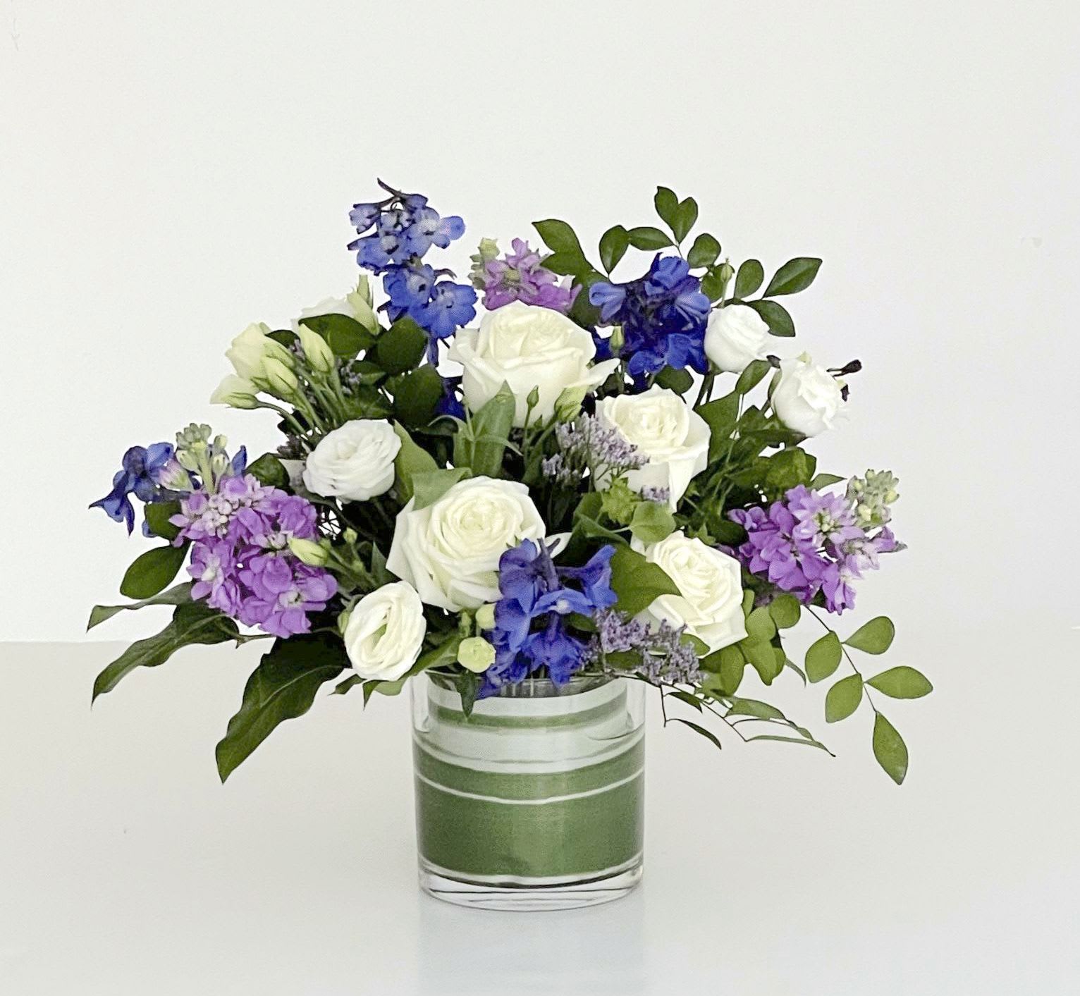 Blue Skies - Market Size - A low leaf-lined cylinder filled with roses, Stock, Lisianthus and Delphinium - a classic and crowd-pleasing color palette. This arrangement is the smallest size we offer of our Blue Skies Collection, but it is still full of flowers. The design is approximately 10" tall and 10" wide - perfect for a dining table, desk, or bedside table - sending a strong message of love, joy, care and celebration through the beauty of flowers.   