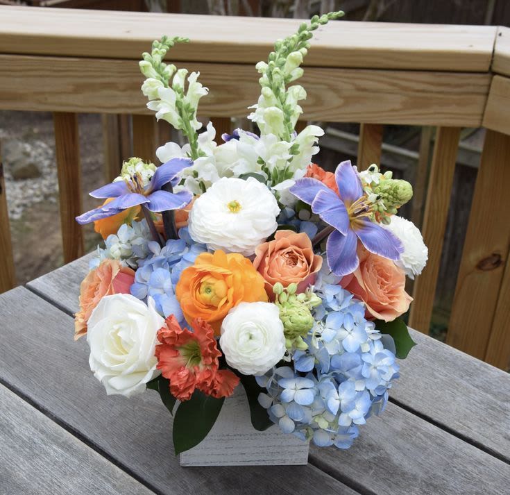 Morning Haze Arrangement - This playful medley features a lush base of billowy blue hydrangeas and creamy white roses, accented by the vibrant, layered petals of orange ranunculus.