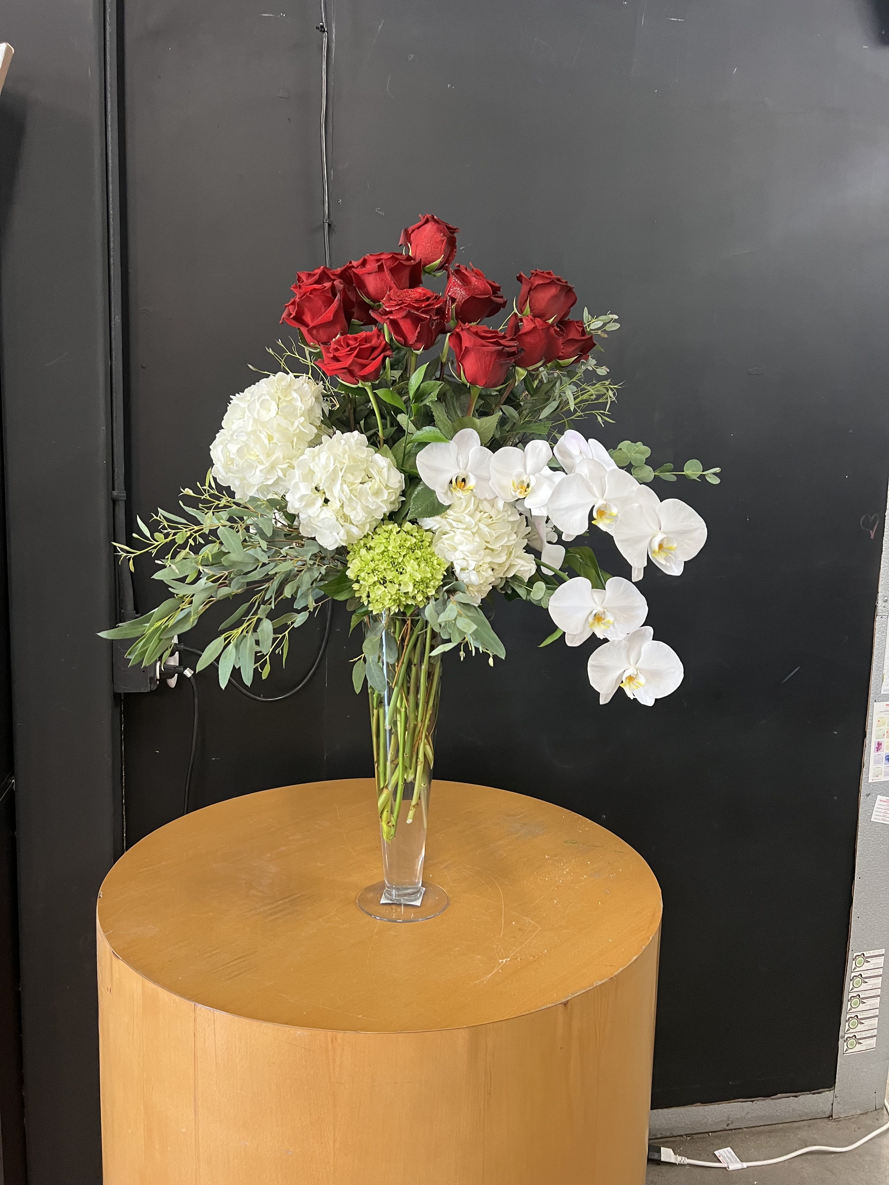 Elegant Roses and Orchids - A striking tall bouquet that pairs classic roses with cascading phalaenopsis orchids for an elegant anniversary or wedding arrangement - the formal red-and-white contrast reads romantic and refined while the airy vertical shape makes a strong visual statement suitable for sympathy or upscale events as well. The person receiving this will feel honored and truly seen: the clustered red rose focal points convey deep affection, while the trailing white orchids add graceful movement and a quiet, luxurious presence.  Carefully composed in a clear glass vase that reveals the long, gathered stems, the design mixes red and white roses, generous white hydrangea clusters, and creamy-white to pale-yellow phalaenopsis orchids. Glossy tropical leaves, palm fronds, and eucalyptus-like greens give height and structure, while mixed pastel and yellow rose groupings can appear in alternate variations for a more celebratory color story. The tall, layered silhouette measures as a statement centerpiece and arrives ready to display - trim stems slightly and refresh the water every two days to keep blooms at their best.