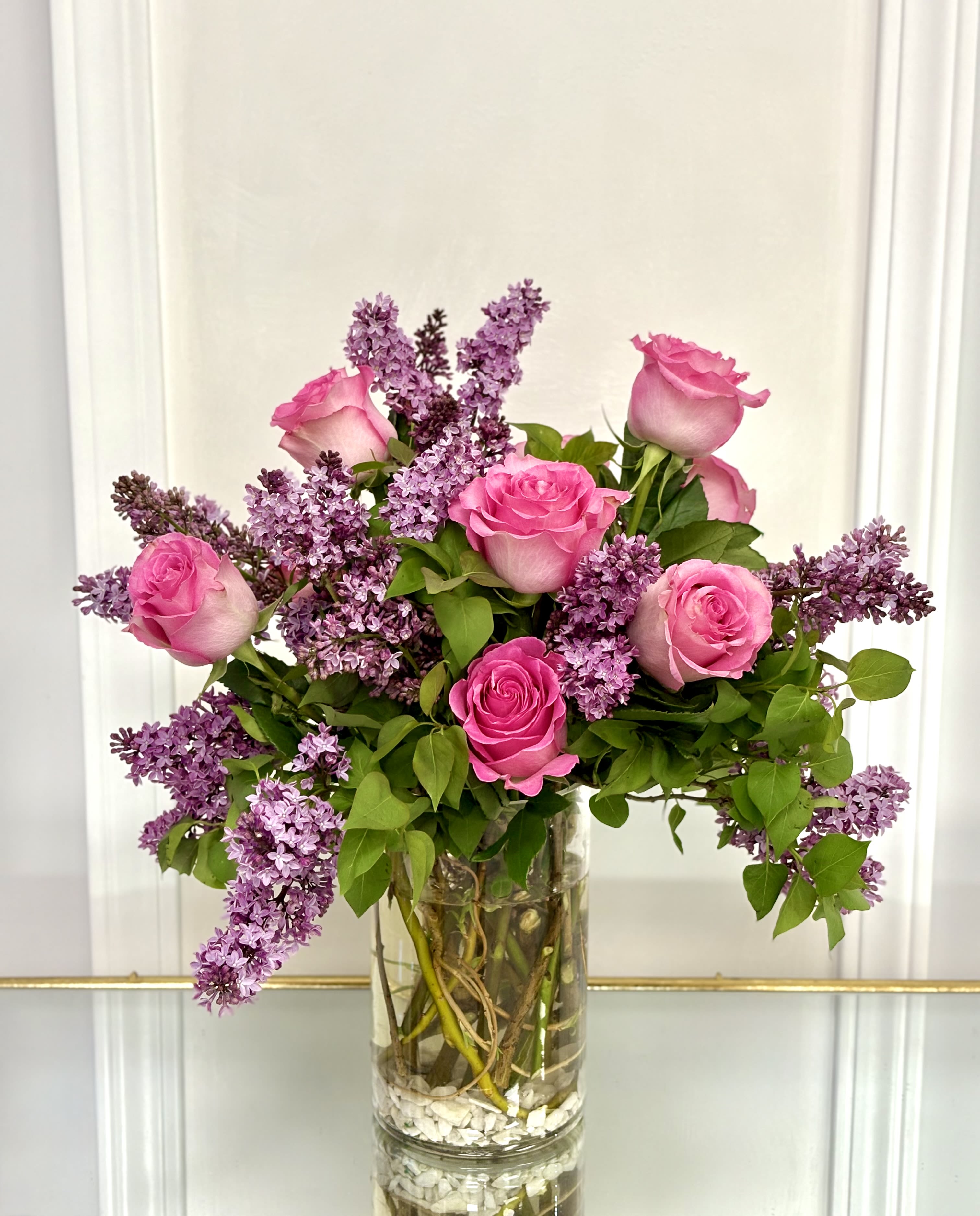 Lilac Bloom Romance By VLM Flowers - A cluster of bright pink roses nestled in fragrant sprays of purple lilac creates an immediate romantic impression-perfect as an anniversary bouquet, a Mother's Day surprise, or a "just because" gift to make someone feel truly seen. The mix of saturated pink and soft lavender reads fresh and garden-inspired, sending a message of love and warmth for birthdays, romantic milestones, or everyday celebrations.  This rounded arrangement is presented in a tall clear glass vase accented with white decorative stones, giving the composition a clean, polished look that highlights stems and water for a crisp modern feel. The combination of lush rose heads and airy lilac adds both visual weight and delicate texture; expect a fullness that fills a room with soft color and gentle scent. Standard vase dimensions and care: keep water fresh, trim stems every few days, and place away from direct heat to enjoy peak bloom life.