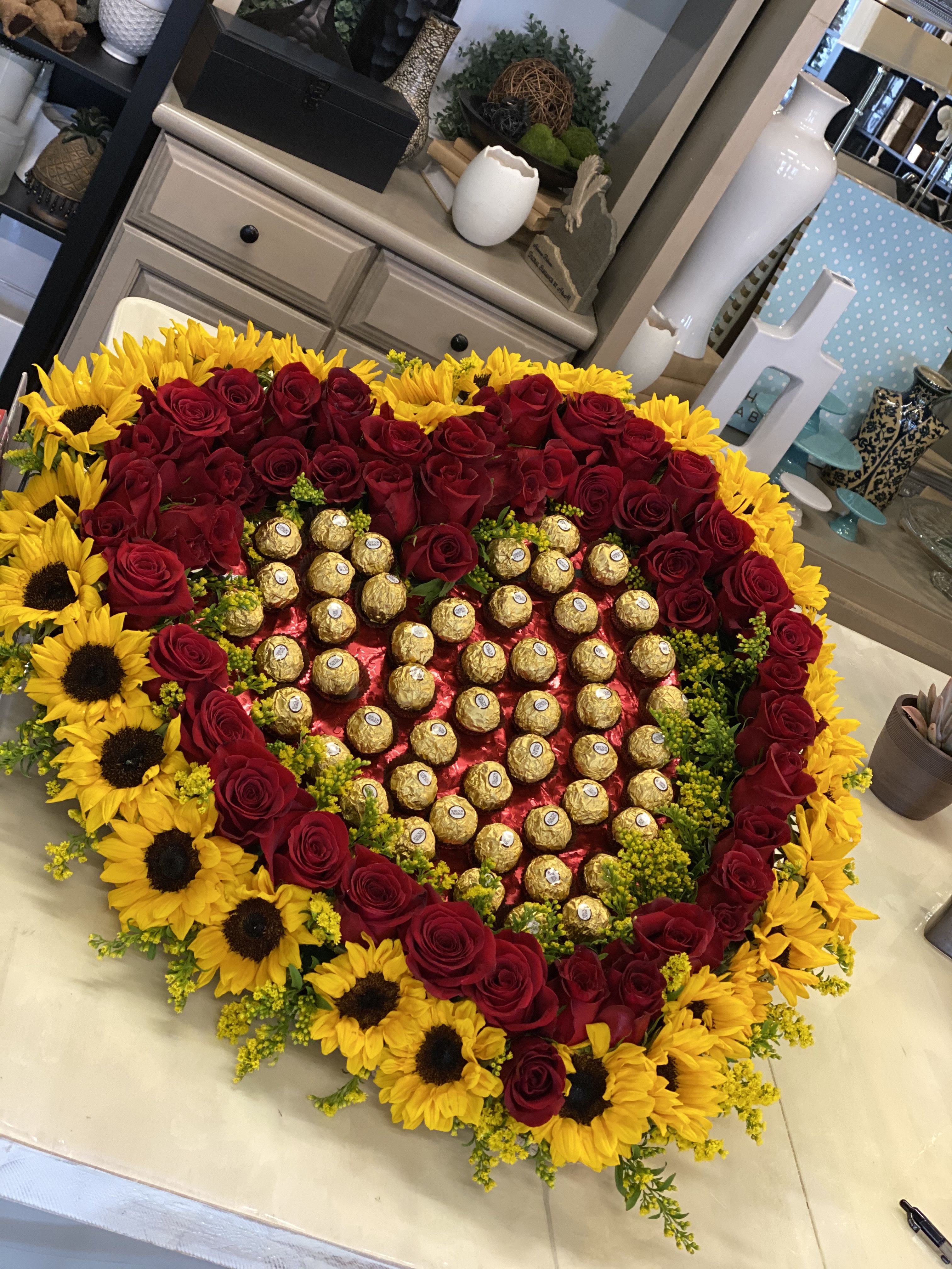 Roses and Sunflower Heart - A bold heart-shaped floral arrangement that reads like a gift: a Roses and Sunflower Heart perfect for an anniversary, Valentine's Day, or any love-themed celebration. The dense border of deep red roses frames vivid yellow sunflowers, creating a dramatic red-and-yellow contrast that feels both romantic and joyfully celebratory-she'll see this as a thoughtful, show-stopping expression of love the moment it arrives.  At the center, individually wrapped chocolates sit on a red, heart-shaped floral base, turning the arrangement into a keepsake-and-treat presentation rather than a traditional bouquet. The texture of velvety rose petals against the sunny, bold faces of the sunflowers adds visual warmth, while the candy centerpiece makes the gift feel personal and indulgent. Size and exact counts follow the pictured large heart format, designed to be a focal gift that fills a room.  This is the kind of arrangement that marks a milestone or makes someone feel truly seen-an anniversary surprise, a Valentine's Day statement, or a romantic "just because" gesture. Order for pickup or delivery to ensure the chocolates and flowers arrive fresh; arrange delivery timing so the recipient enjoys both the bloom and the treats at their peak.
