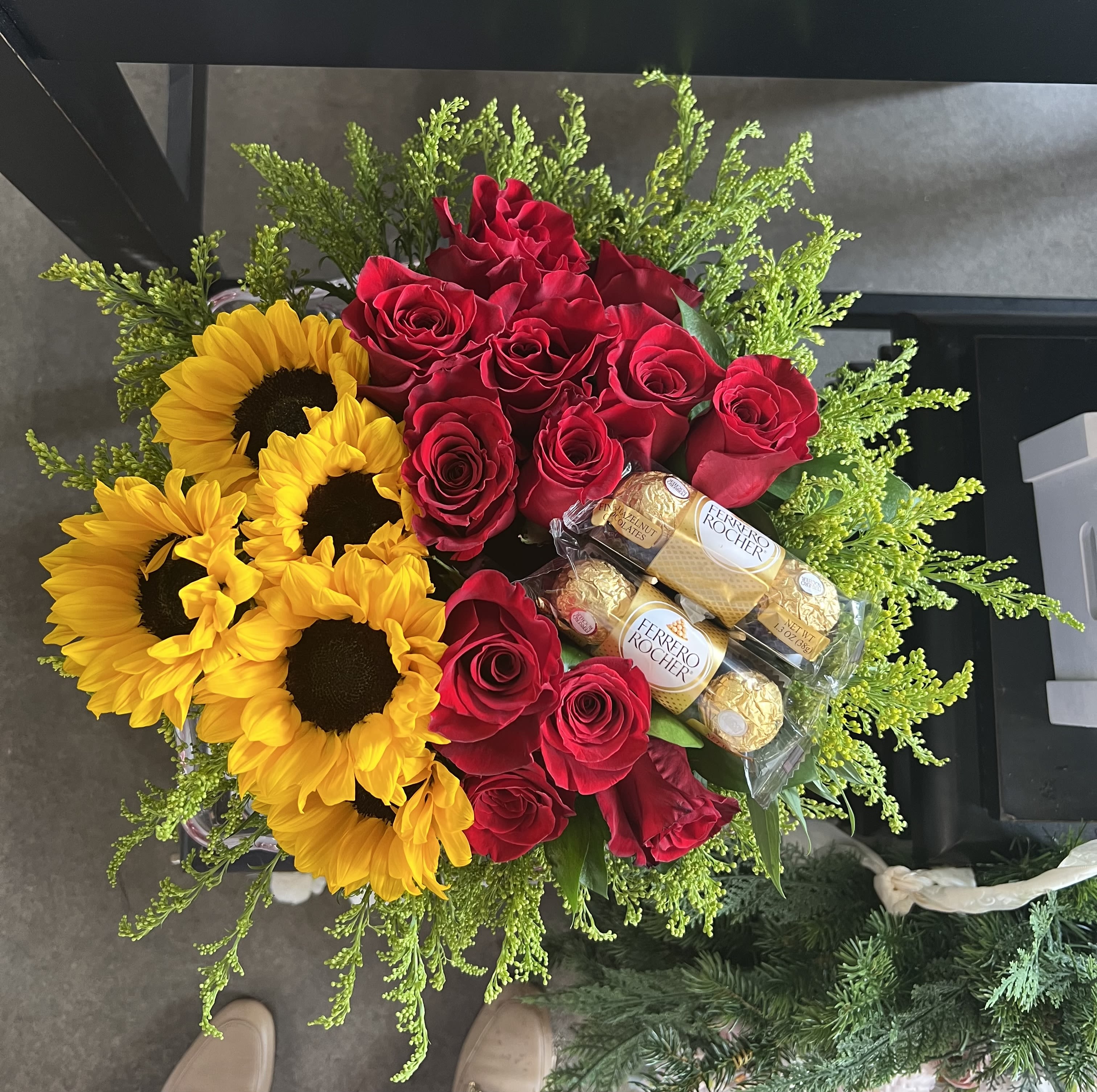 Roses and Sunflowers Forever - A bold red-and-gold contrast greets the recipient: clustered red roses paired with striking yellow sunflowers make this flower arrangement a joyful choice for an anniversary, birthday, or Valentine's Day bouquet. The mix reads romantic and celebratory at once-she'll feel truly seen by the warmth of the sunflowers and the classic romance of the roses.  The arrangement is shaped into a full, rounded bouquet with airy green filler that frames and lifts the blooms, giving a lively, fresh silhouette. Ferrero Rocher chocolates are tucked into the design, wrapped in clear plastic for a gift-ready presentation that combines flowers and sweets in one elegant package. Colors include rich red, sunny yellow and lively green, producing a vivid, festive look that photographs beautifully.  Perfect for marking romantic milestones or surprising the person you're thinking of on their birthday, this bouquet arrives ready to gift and makes an immediate impression. For best enjoyment, keep the stems in fresh water and away from direct heat or drafts to extend the life of the roses and sunflowers.