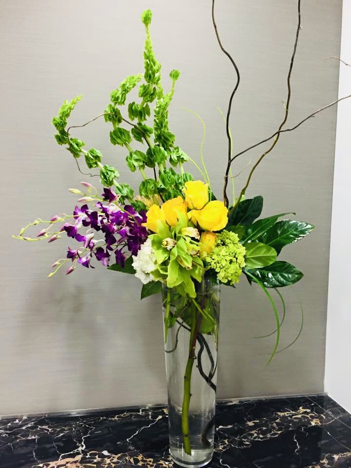 Modern Elegance - Sunlit yellow roses paired with vibrant purple orchids create a bold, celebratory bouquet-perfect as a birthday bouquet, congratulatory arrangement, or upscale everyday gift. The vertical, sculptural silhouette feels dramatic and modern: looping curly branches arc above the blooms so the person you're thinking of will first notice height and movement, then the rich color contrast that makes an entrance feel festive.  This tall arrangement uses yellow roses, purple orchid sprays, green-toned blooms (including hydrangea-like texture), and crisp white accents arranged in a clear cylindrical glass vase so the layered stems remain part of the visual interest. Glossy foliage and decorative curly branches give the design an airy, tropical feel while keeping the focus on saturated color and clean lines.  Ideal for celebrating milestones, marking a promotion, or dressing a table for a formal event, this composition reads as both lively and refined-she'll feel genuinely seen when these blooms arrive. The clear glass vase emphasizes stem length and verticality; arrange on a console or pedestal where the height can be appreciated, and refresh water every 2-3 days to keep blooms looking their best.