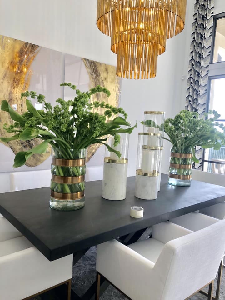 Bells of Ireland - Lush, architectural stalks of green create an immediate sense of modern elegance-this Bells of Ireland flower arrangement is ideal as a centerpiece for a formal dinner, rehearsal dinner, or green-and-white anniversary celebration. The tall, airy profile and vivid green tones draw the eye upward, so the person you're honoring will feel noticed and admired without the arrangement blocking conversation across the table.  Arranged in clear glass vases with a subtle gold band, the composition pairs clustered green blooms and broad glossy leaves for a fresh, sculptural look. The design's vertical lines and open negative space give it a contemporary, refined presence on a dark dining table; shown with white cylindrical candle holders and a chandelier-lit setting, it complements upscale event decor and clean, minimalist interiors.  Perfect for a recipient who prefers understated luxury, this piece works as a statement centerpiece or an elegant room accent that reads both celebratory and serene. Vases are tall to showcase the arrangement's height-place on a large dining table or buffet; swap in battery candles or low-profile lighting for long-lasting ambiance. Order in advance for event delivery to ensure peak freshness.