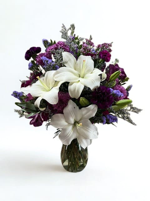 Petals of Remembrance  - White lilies take center stage in this sympathy-leaning flower arrangement, making it a quiet, elegant choice for a condolence or remembrance bouquet. As a flower arrangement suited for memorial services or to send your thoughts, the mix of white, purple, and pink evokes calm dignity while still feeling gentle and personal - the kind of gift that helps someone feel seen in a difficult moment.  The composition is full and rounded, with large white lilies opening prominently at the front and taller stems rising slightly above a dense cluster of purple and magenta blooms, accented by small lavender filler. Carnations and other complementary flowers add soft texture and depth, all arranged in a clear glass vase that lets the natural stems show and fits comfortably on a mantel or small table. The palette reads formal without being austere, blending crisp white with saturated purples and pinks for a composed, soothing presence.  Perfect for sending sympathy, expressing condolences, or honoring a life remembered, this arrangement offers a respectful balance of grace and tenderness for the person you're thinking of. It arrives ready to display in the included clear glass vase; to keep it looking fresh, change the water every couple of days and trim the stems slightly when refilling.  As similar as possible. Designer may make appropriate changes based on flower availability. 