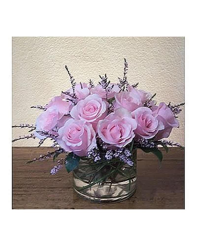 Short arrangement of light pink roses with purple filler in a glass vase