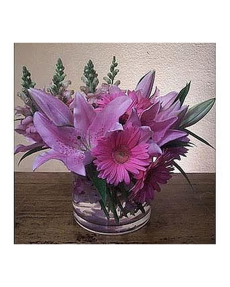 Low arrangement of pink lilies and gerbera daisies in a clear glass vase on a wooden surface