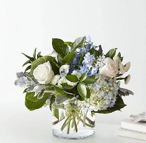 Short arrangement of white roses and blue hydrangeas in a clear glass cylinder vase