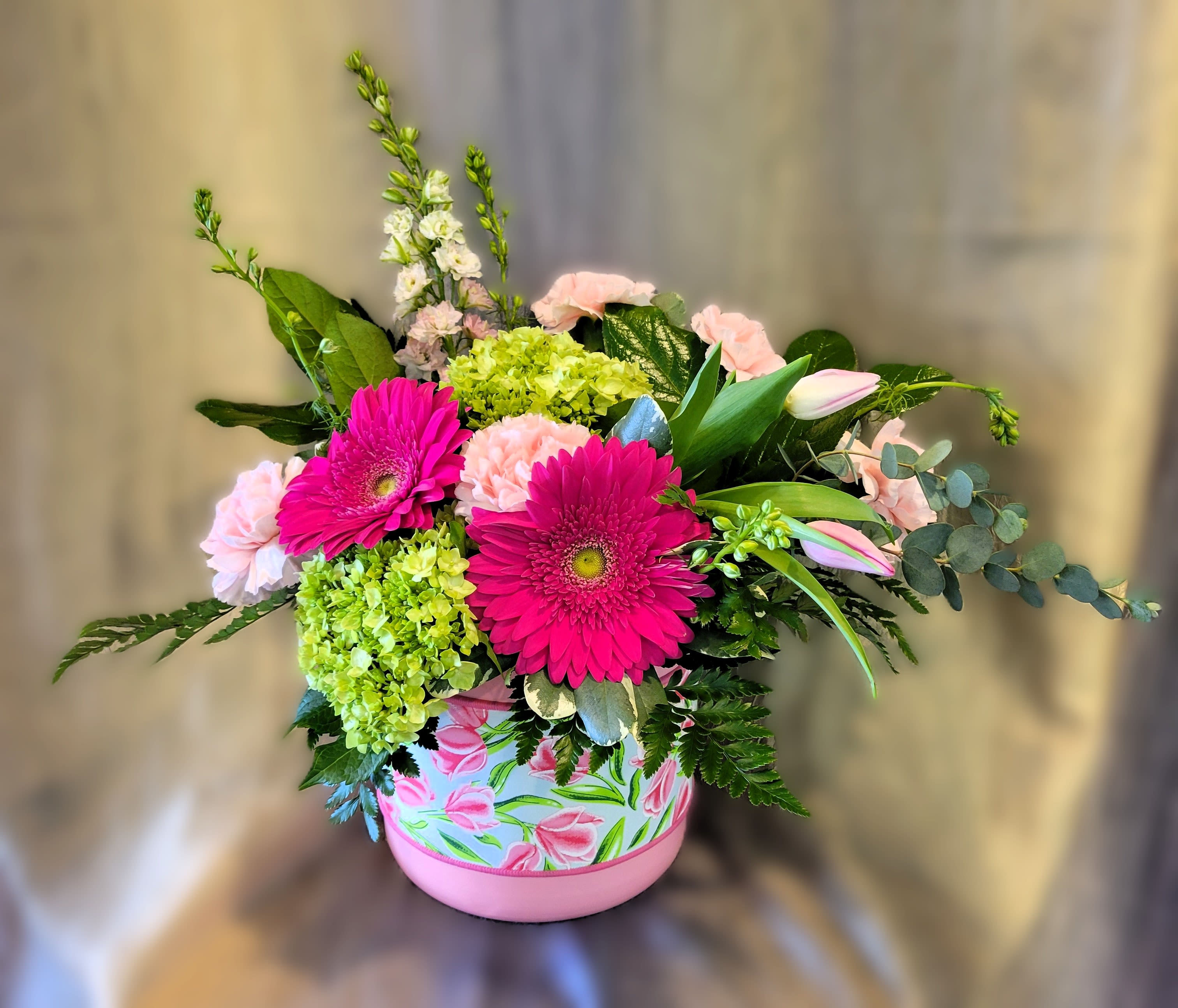 Rhapsody ~ Flower A Day - Hot-pink gerbera daisies take center stage in this lively flower arrangement, making it a joyful choice for a Mother's Day bouquet, anniversary gift, or Birthday surprise. The combination of bright pinks and soft pastels-pink carnations, lime-green hydrangea, tulips, purple accents, and larkspur-creates an energetic, garden-fresh look that reads as both playful and elegant. Designed to delight the person you're thinking of, it sends a cheerful, uplifting message perfect for Thank You, Get Well, Housewarming, Retirement, or romantic moments.  This full, slightly asymmetrical design spills outward with varied textures: the smooth gerbera faces, ruffled carnation petals, clustered hydrangea, and tall larkspur spikes for height and movement. Arranged in a decorative round container wrapped with a floral-print ribbon, the piece has a feminine, modern feel that fits a bedside table, office desk, or entry console. Size and exact stem counts vary by season; flowers may be substituted with similar varieties to preserve color and overall style, ensuring the recipient still receives a fresh, thoughtfully composed arrangement that makes them feel truly celebrated.