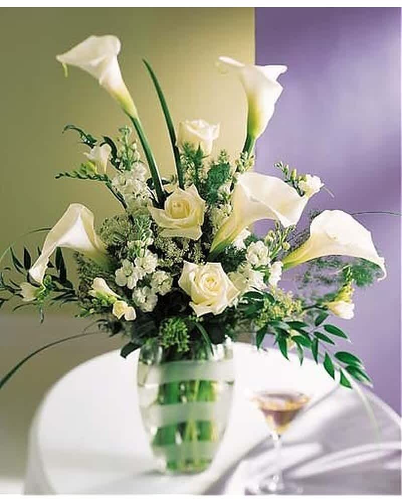 Tall arrangement of white calla lilies and roses in a clear glass vase with green ribbon accents