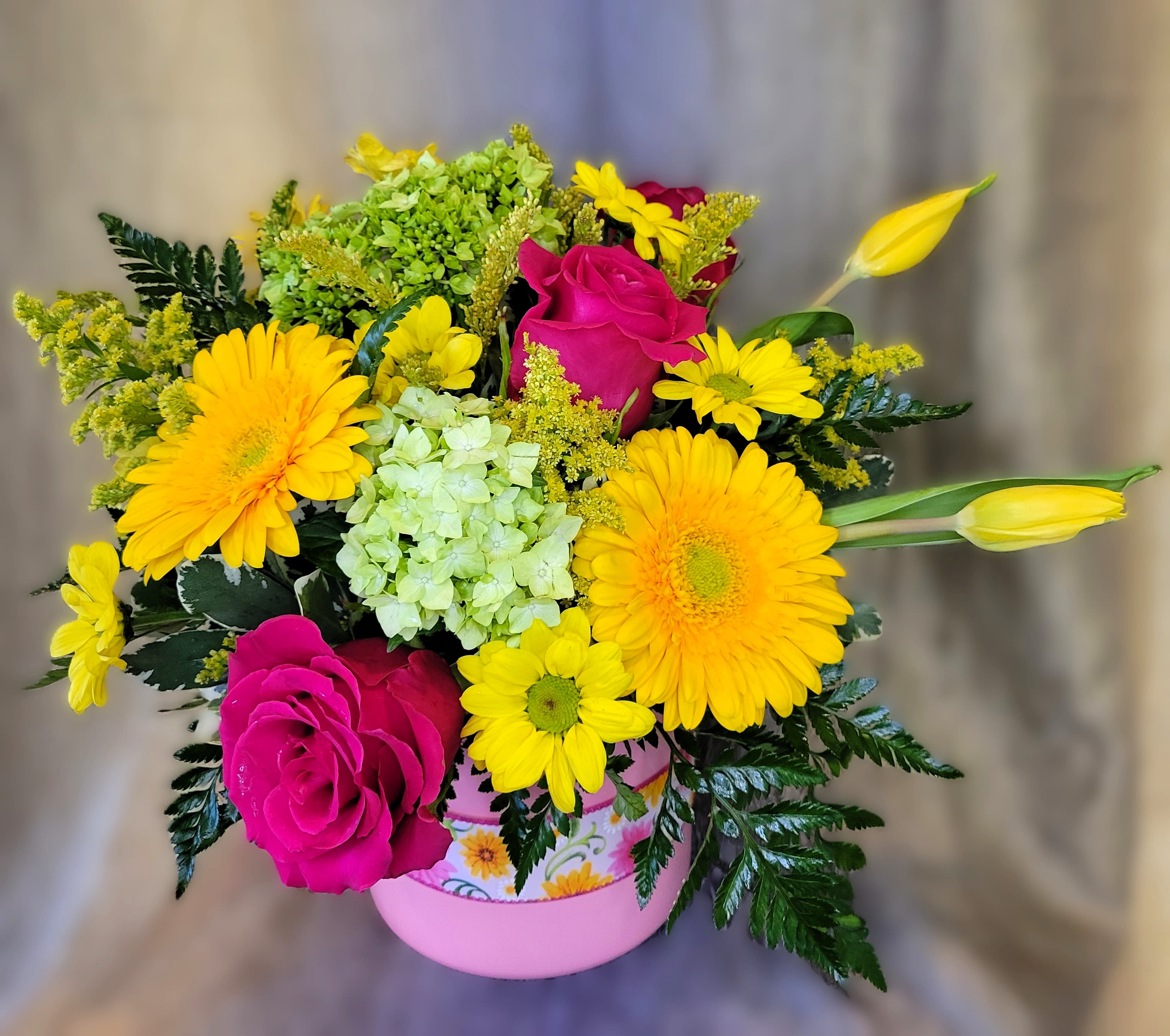 Mama's Garden Whimsy ~ Flower A Day  - Bright hot-pink roses and sunny yellow gerbera daisies meet pale green hydrangea and yellow daisies in a lively spring bouquet designed for Mother's Day, birthdays, thank-you gestures, or "just because" moments. This flower arrangement for gifting places the occasion front and center: it's the kind of cheerful birthday bouquet or get-well pick-me-up that makes the person you're thinking of feel seen and genuinely celebrated.  The rounded, full composition pairs contrasting colors-vivid pinks, clear yellows, fresh greens and touches of white-for maximum visual impact. Yellow tulip buds extend outward to add movement, while mixed daisies and pale green hydrangea provide texture and volume around the roses. The stems arrive arranged in a pink vessel with a floral pattern that complements the palette and ready to display.  Ideal for moms, friends, or coworkers who appreciate bright, upbeat florals, this arrangement suits springtime events or any day you want to lift someone's spirits. Sizes are arranged to create a full, rounded silhouette; to keep blooms looking their best water daily with cold water.
