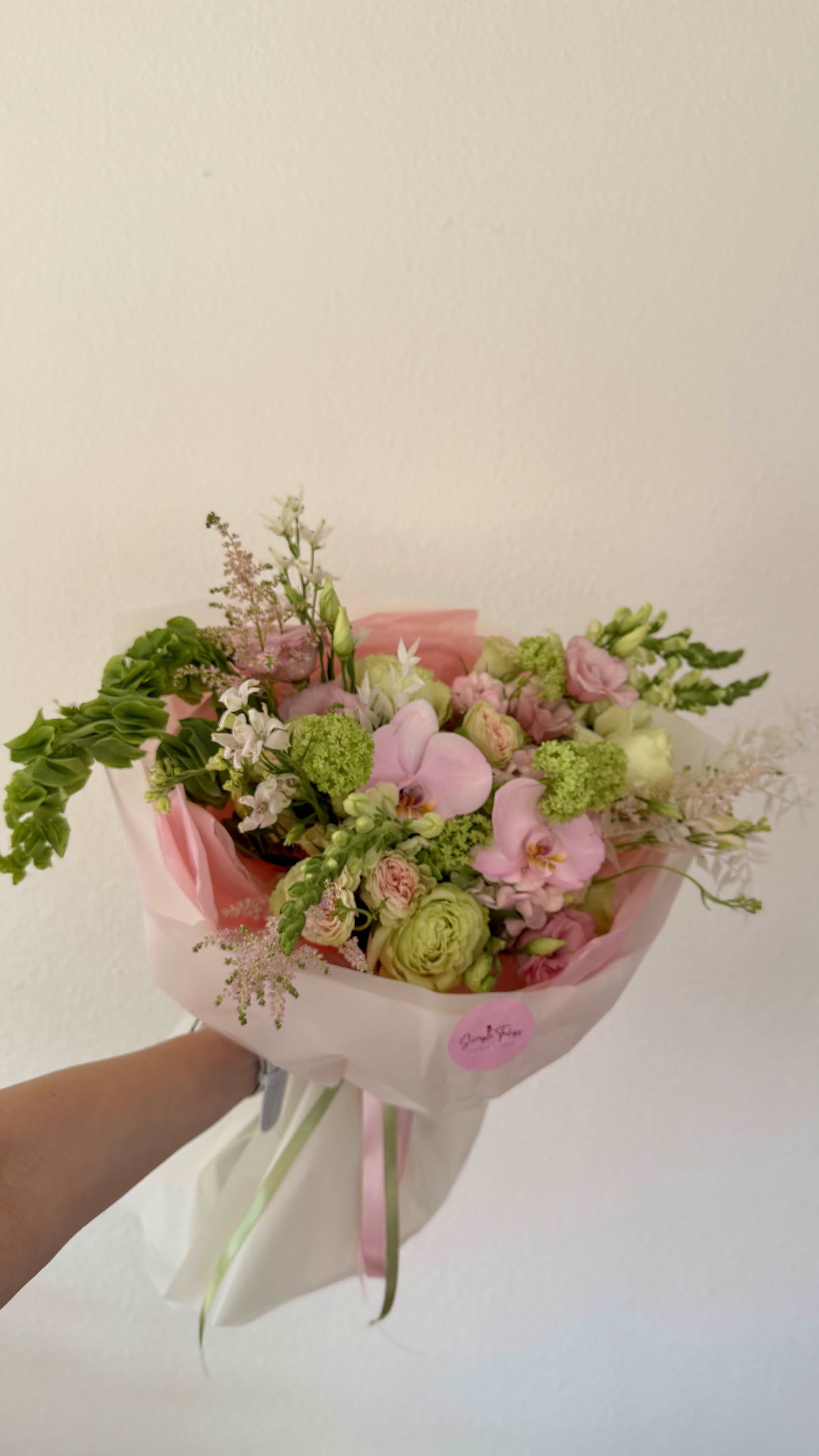 Strawberry Matcha Bouquet - Soft pink phalaenopsis orchids set a romantic tone in this birthday or anniversary flower arrangement, their elegant blooms paired with pale green accents for a fresh, springtime feel. The Strawberry Matcha Bouquet reads cheerful and garden-like-perfect for the person you're celebrating when you want them to feel truly seen and delighted.  This hand-tied bouquet mixes clustered roses, ranunculus, lisianthus, and daisies with airy white accents and delicate filler for a textured, abundant look. Wrapped in pink tissue and white paper with long ribbon tails and a round sticker label, it arrives gift-ready and polished. The palette of pink, green, white, and hints of purple creates soft contrast and visual depth while the orchids provide graceful height and focal interest.  Ideal for birthdays, anniversaries, or any spring celebration, this bouquet feels both luxurious and effortless on the recipient's table. Arrange in a vase with fresh water on delivery day; trim stems and refresh water every other day to help blooms last longer.