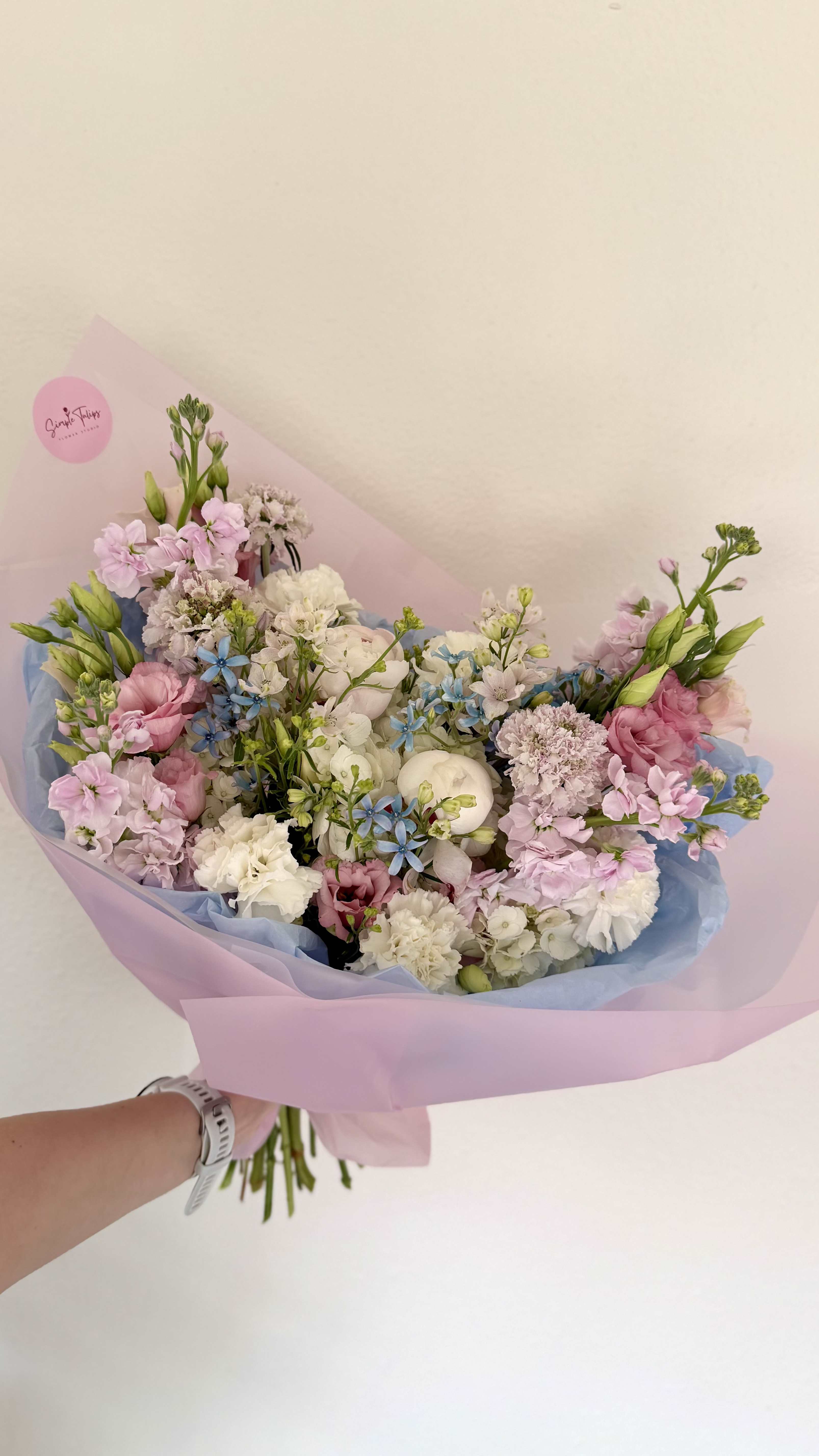 Gentle bloom - Soft pastel blooms gathered in a loose, hand-tied shape-this flower arrangement is a gentle birthday or anniversary bouquet that reads as quietly joyful. The mix of pink, white, lavender and pale blue flowers-featuring ruffled carnations, lisianthus-like blooms, dianthus textures and full white peony focal flowers-creates a soft, romantic impression that will make the person you're thinking of feel seen and celebrated.  Wrapped in layered pink and blue paper for a gift-ready finish, the bouquet's airy, full silhouette is punctuated by small blue star-shaped accent flowers that add delicate contrast and movement. The composition is designed to feel fresh and feminine, perfect for spring gifting or any gentle celebratory moment; choose this arrangement to convey affection, congratulations, or warm thinking-of-you wishes with an elegant, effortless look.